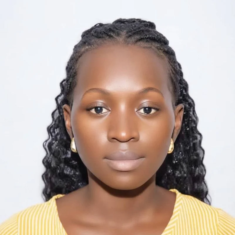 Portrait of a young woman with dark, curly hair, wearing gold earrings and a yellow striped top, standing against a plain white background.