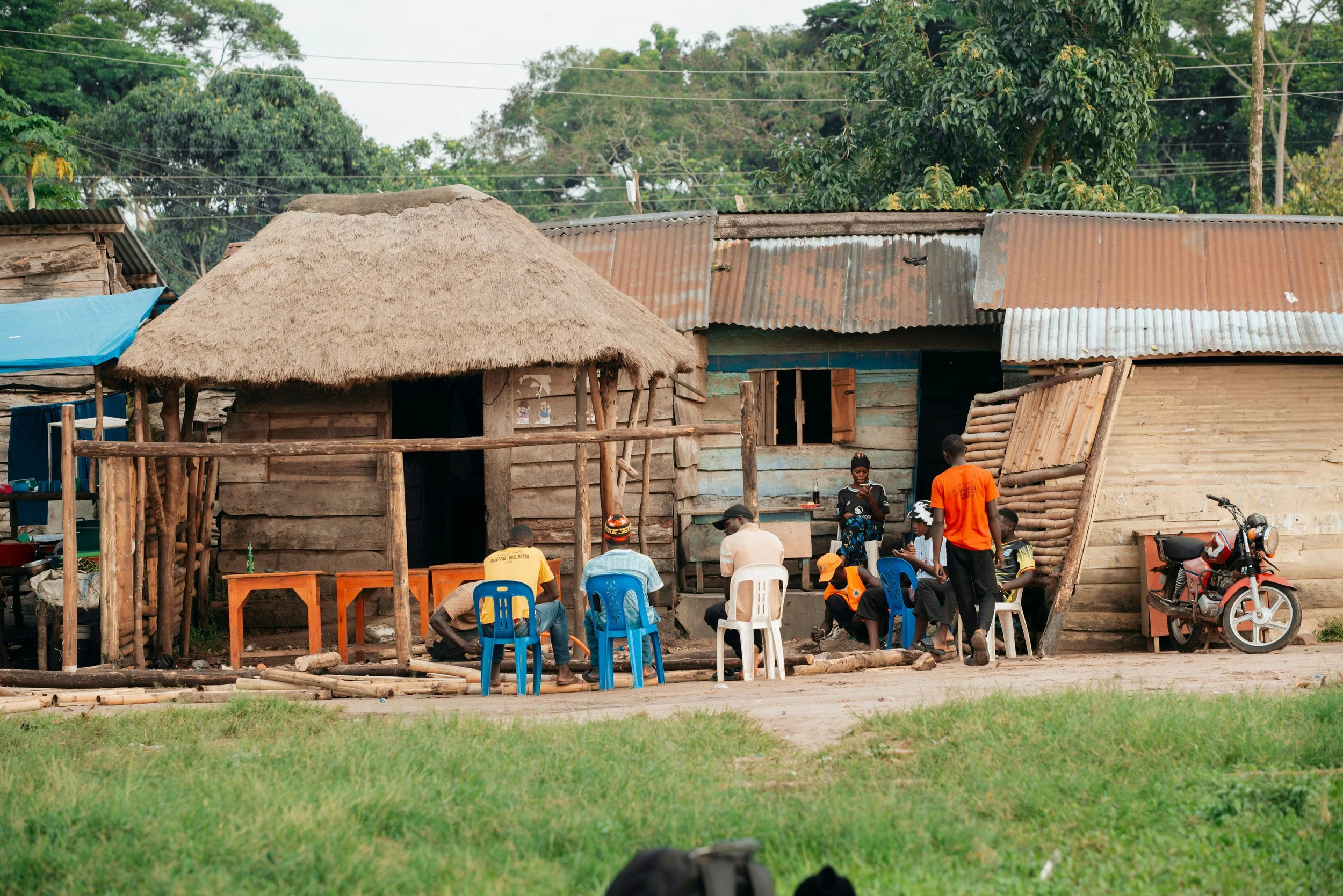 A group of people gathered outside a rustic wooden house with a thatched roof and corrugated metal roofing in a rural area, with trees and power lines in the background.