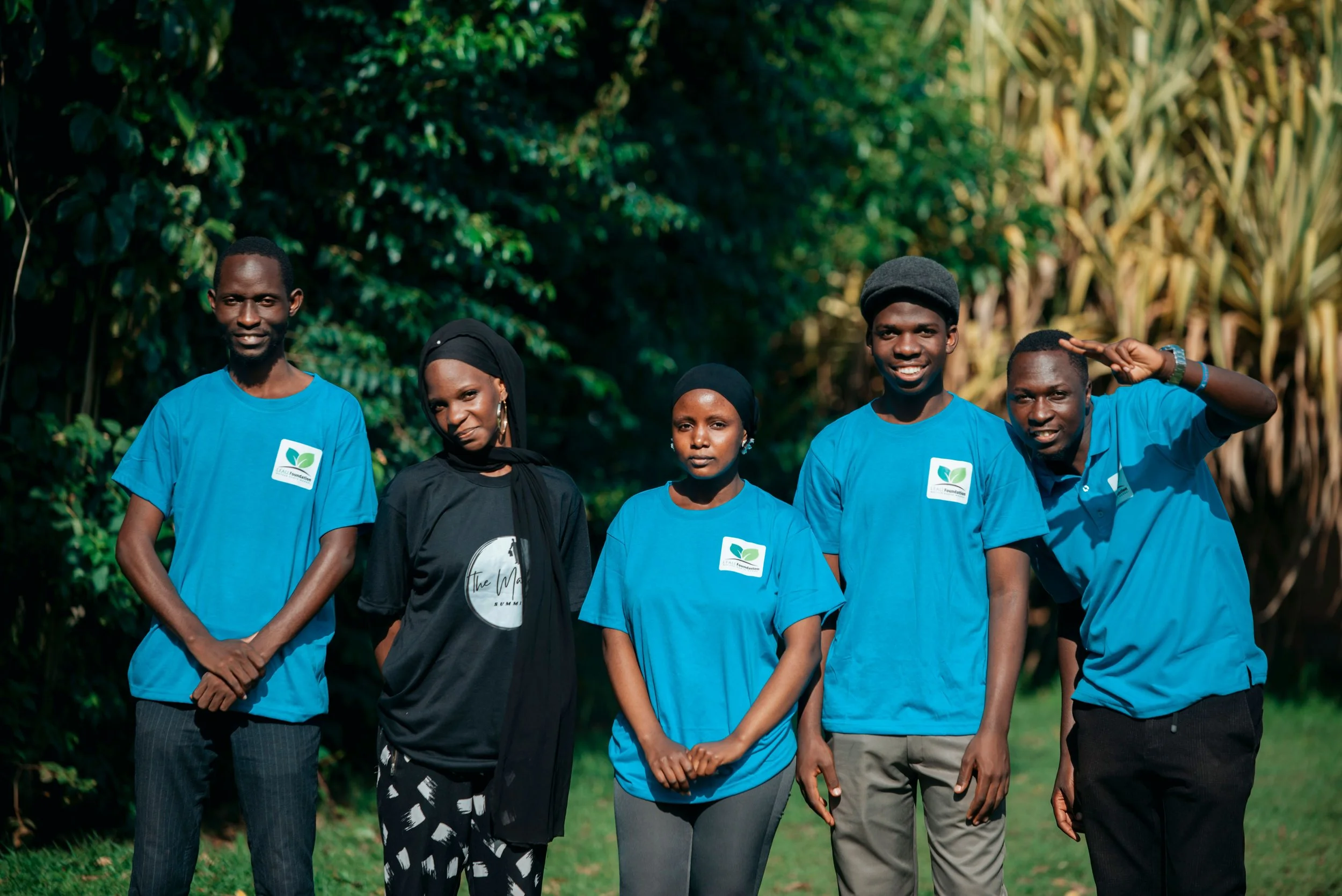 Group of five people standing outdoors in a green, leafy setting, wearing blue shirts with a logo, smiling, and posing for the photo.