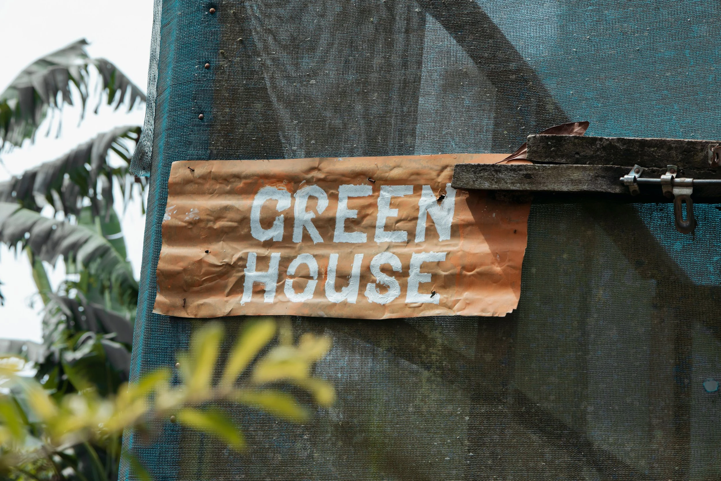 A close-up photo of a sign that reads 'GREEN HOUSE' attached to a textured black surface with a tarp or fabric cover, with green plants and foliage in the background.