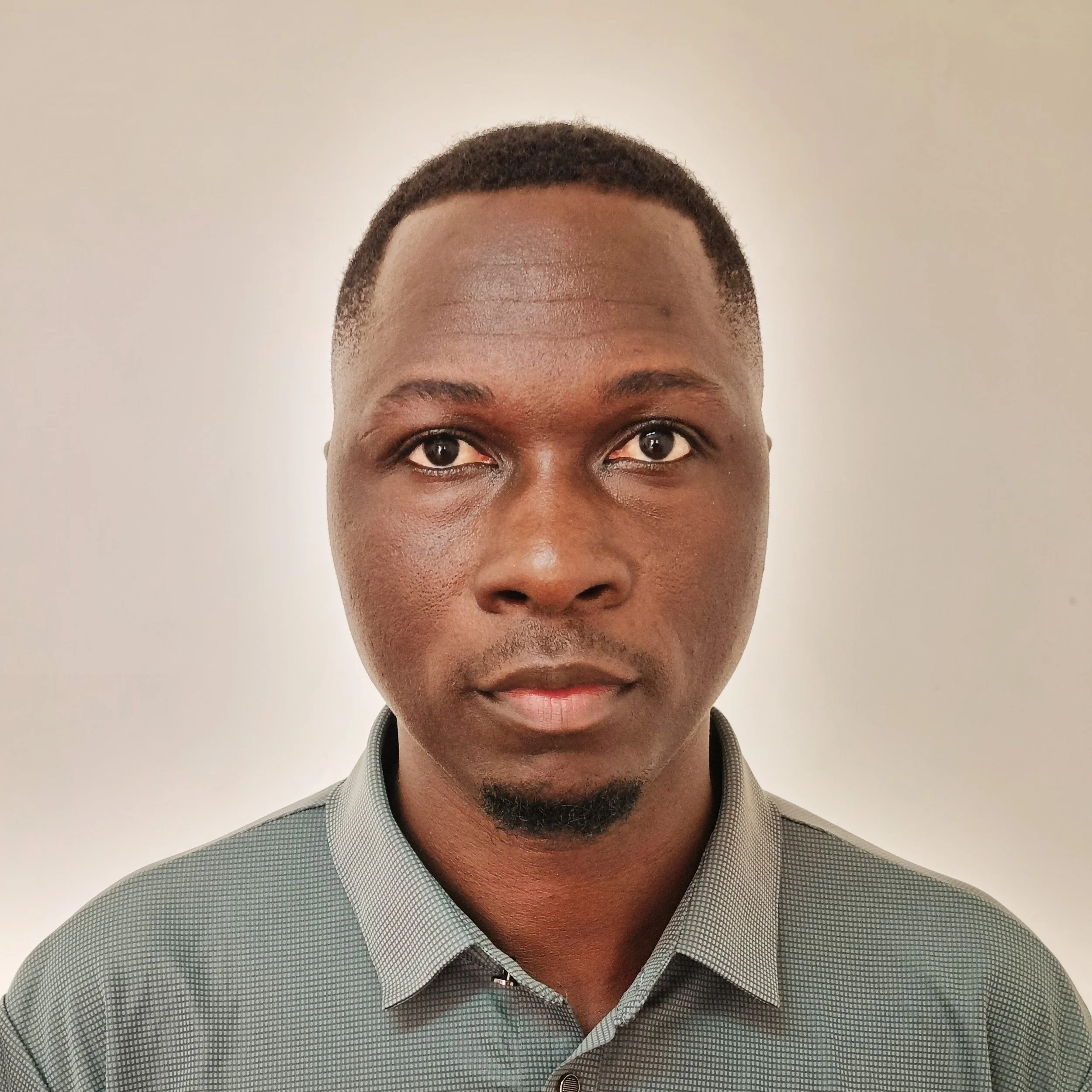 Close-up photo of a man's face and upper body, wearing a gray checked shirt, against a plain beige background.