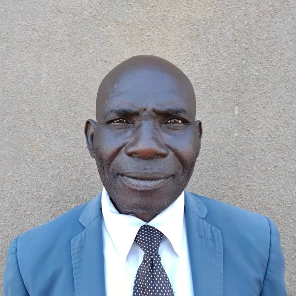 A formal portrait of a bald African American man wearing a blue suit, white shirt, and a black polka dot tie, standing against a beige wall.