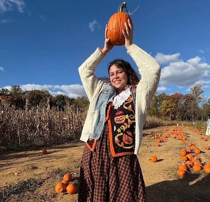 A woman holding a pumpkin above her head in a pumpkin patch during fall, with a blue sky and scattered clouds in the background.