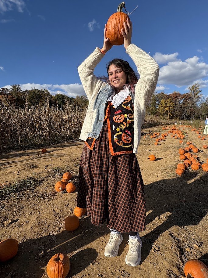 Woman smiling and holding a pumpkin above her head in a pumpkin patch on a sunny day with blue sky and scattered clouds.