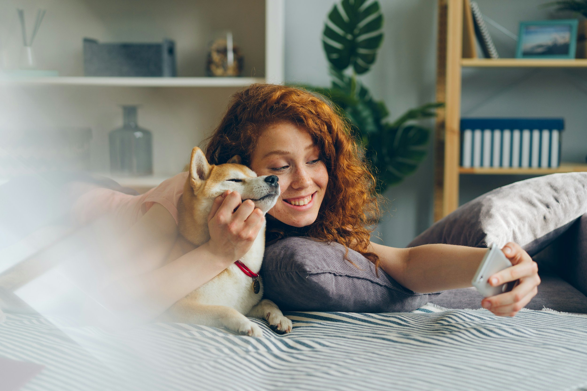 A young woman with curly red hair lying on a sofa, smiling and holding a smartphone, with her dog resting its head on her shoulder.