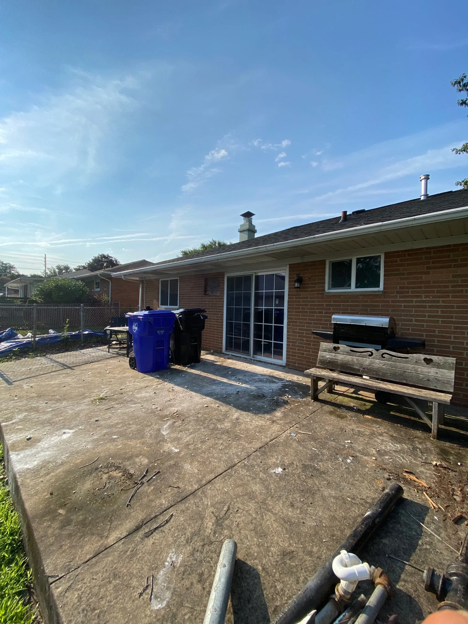 Backyard with a brick house, sliding glass door, and a wooden bench, with blue trash bins and various pipes and debris on concrete ground  H&H property services in Parma Ohio