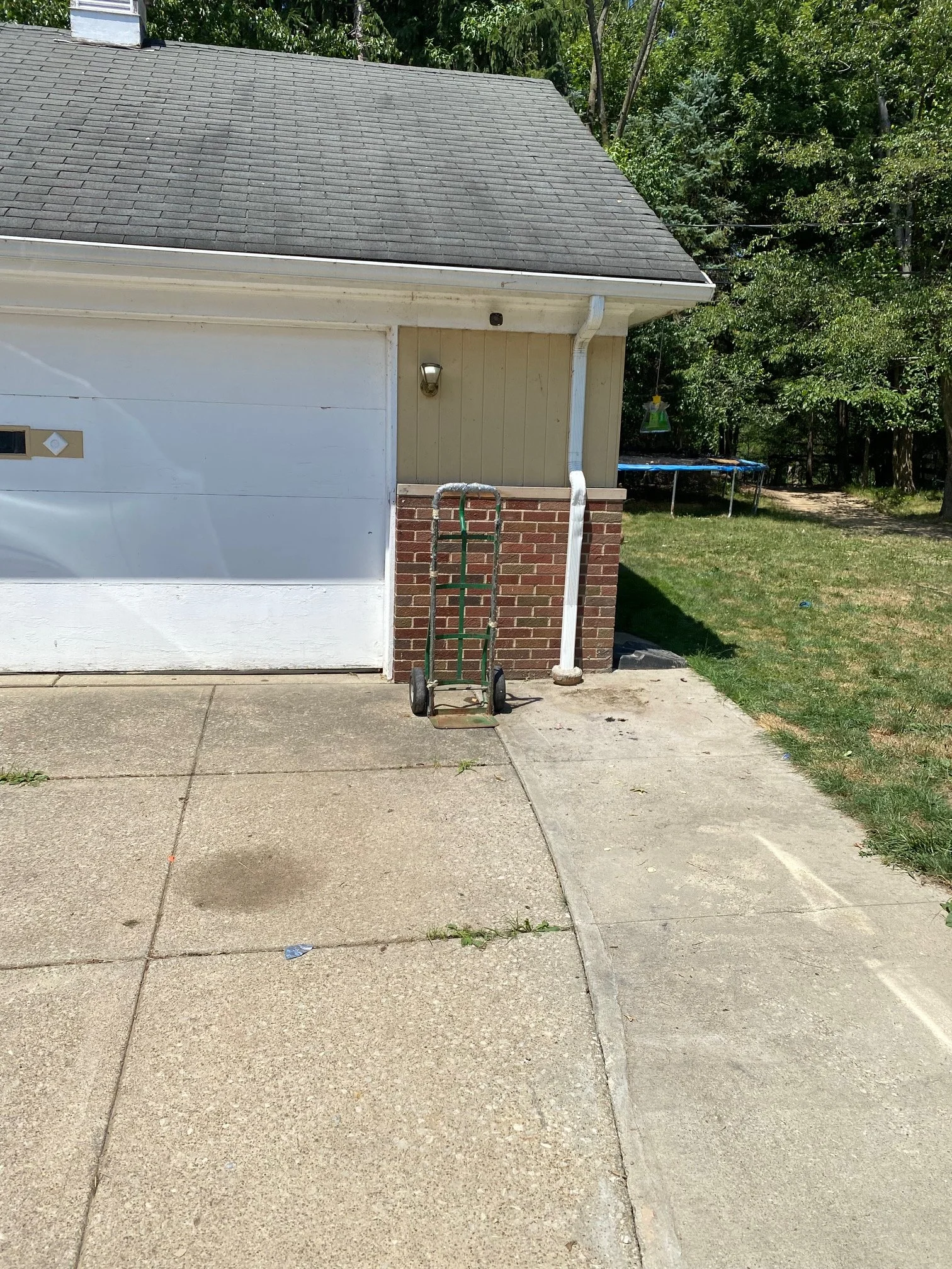 Side view of a house with a gray shingled roof, beige vertical siding, a brick accent, and a white garage door. A hand truck is in front of the house, on a concrete driveway next to a grassy yard with a trampoline and trees.