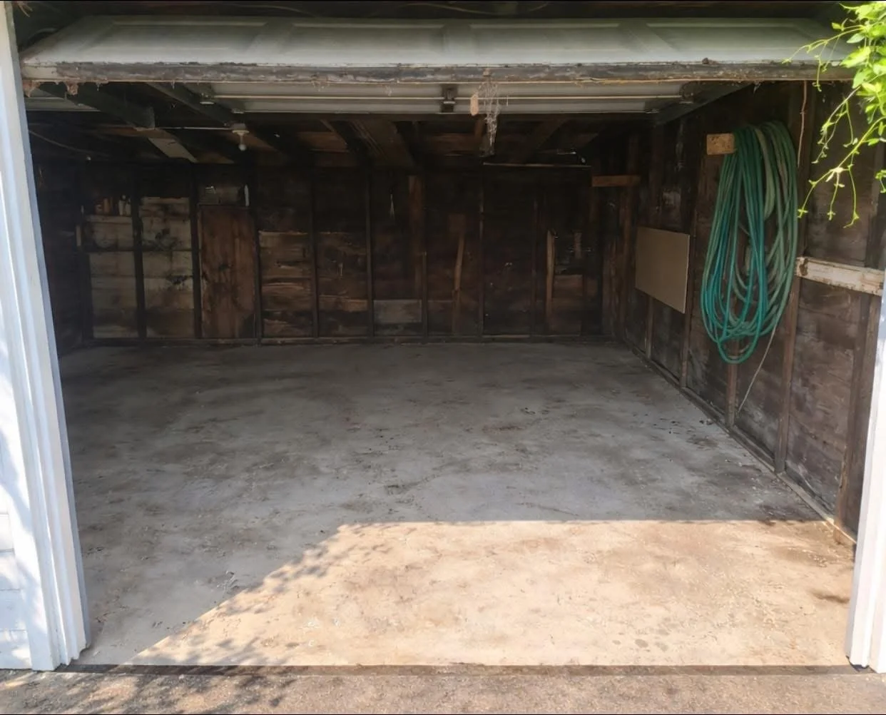 Empty garage with unfinished wood walls and a concrete floor. Green garden hose is coiled on the right wall.