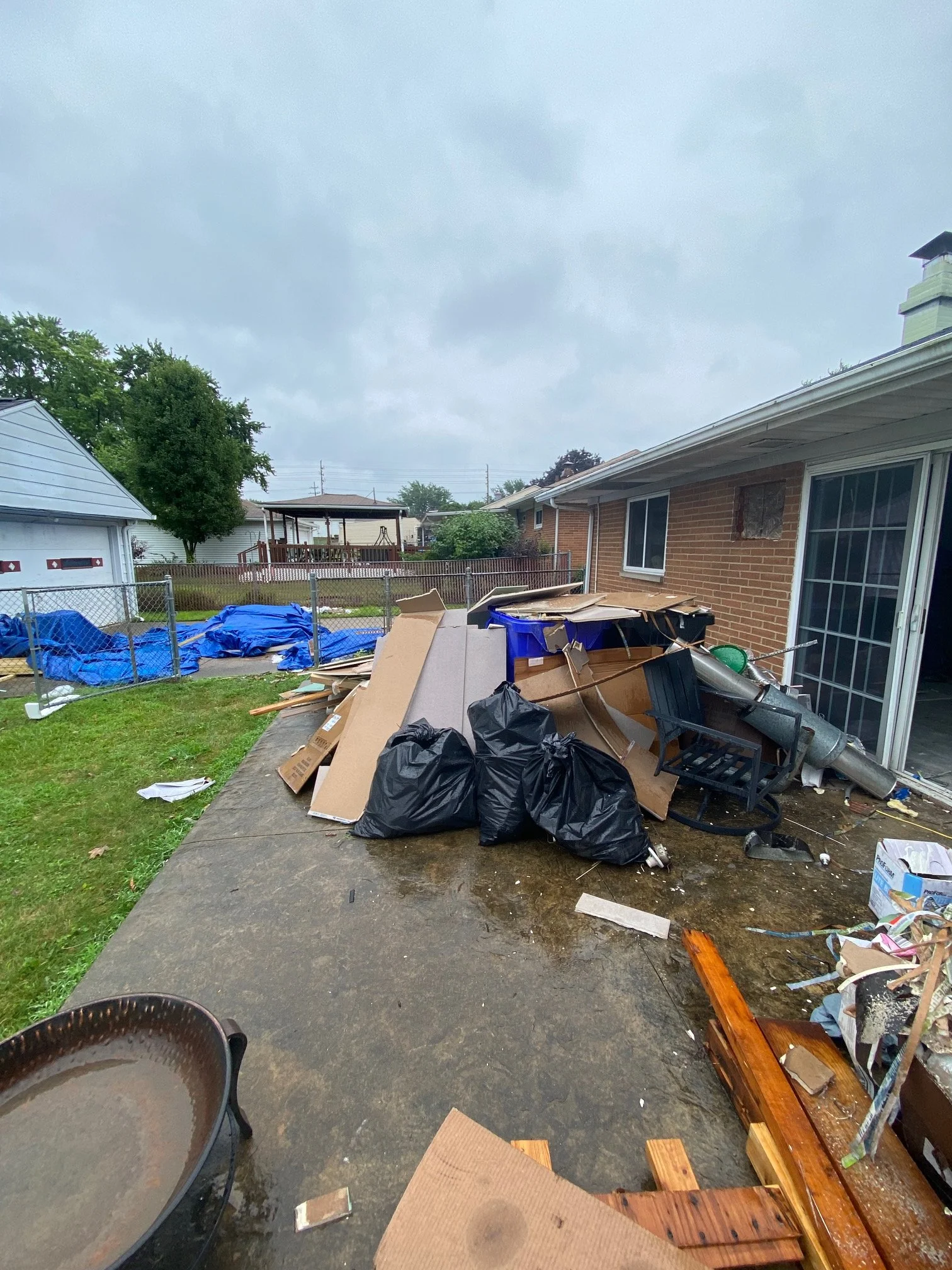 Backyard with scattered debris, cardboard, trash bags, and a black chair. A fence separates the yard from neighboring houses. Overcast sky.