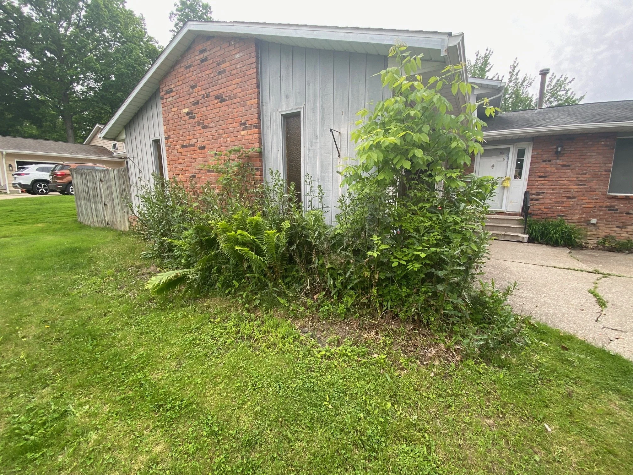 View of a suburban house with a brick and wood exterior, front steps, and a lush, green front yard with plants.