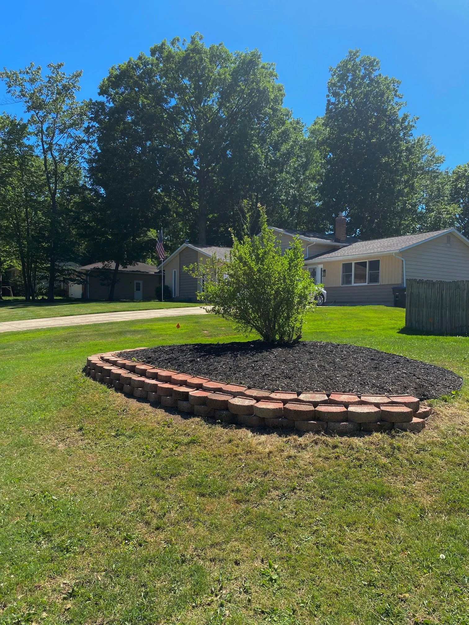 A front yard with a small landscaped area bordered by red bricks, featuring a single small green bush. In the background, there are two houses with light-colored siding, a large tree, a flagpole with an American flag, Brunswick ohio