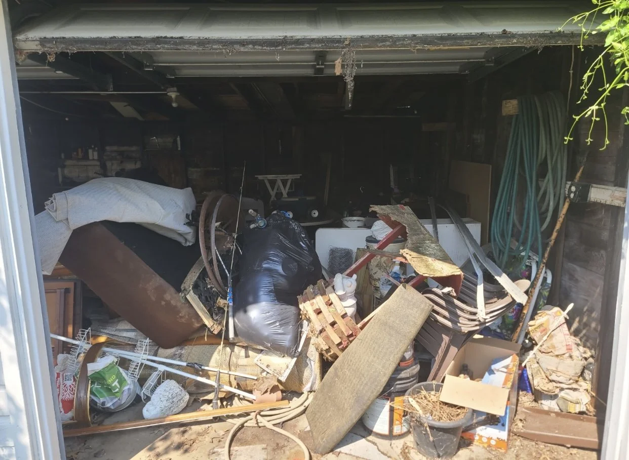 Cluttered garage filled with various household items, furniture, bags, and miscellaneous debris.