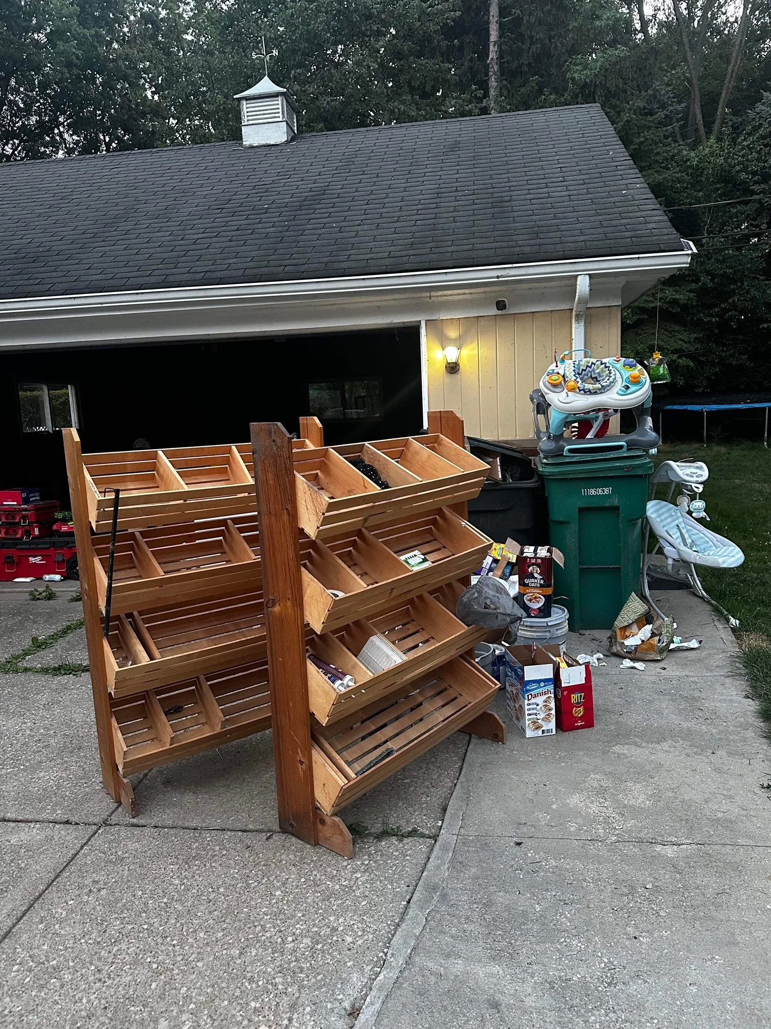A backyard scene with a wooden plant stand in the foreground, a house with beige siding, a sharply pitched dark shingled roof, a weather vane on the roof, a wall-mounted light, and various items including a baby walker, trash cans, and boxes scattere