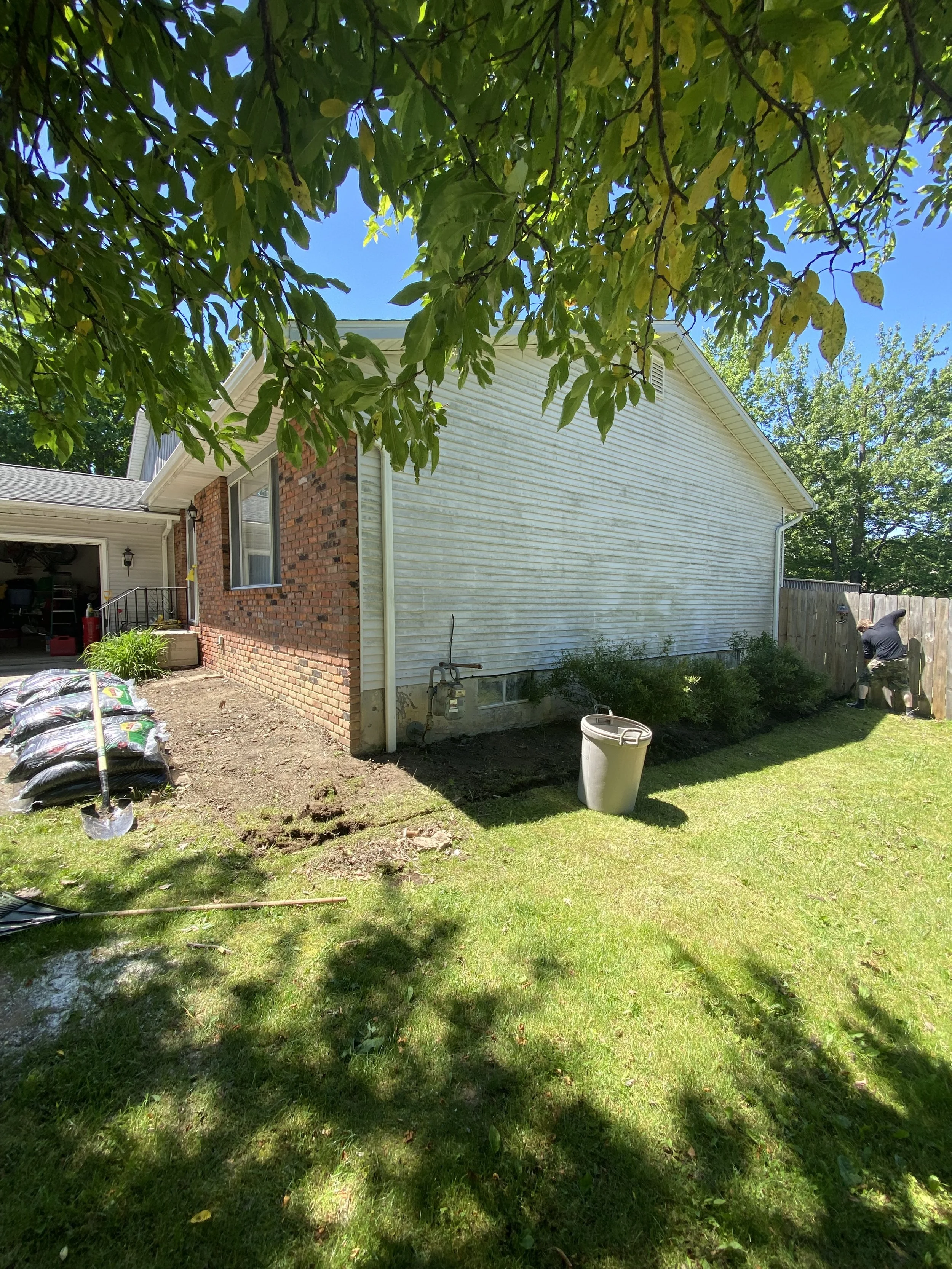 A backyard scene with a house, trees, and a man working near a wooden fence under a blue sky. Gardening supplies and tools are visible near the house. Cleveland Ohio