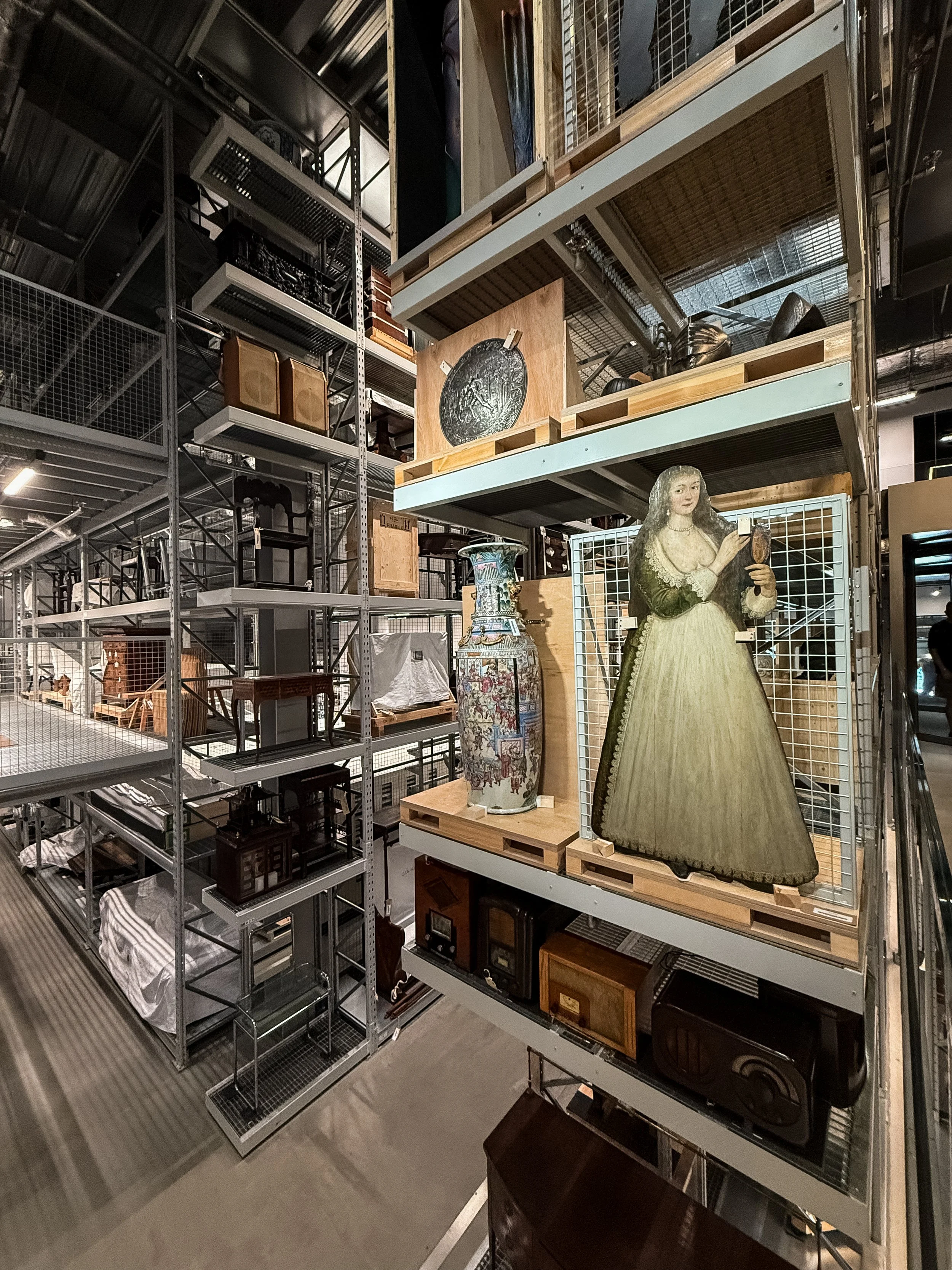 Interior of the V&A East Storehouse museum in London, United Kingdom. Warehouse shelving filled with vintage furniture and decor pieces, including a portrait of a woman in a yellow dress, large porcelain vase, and various boxes and objects.