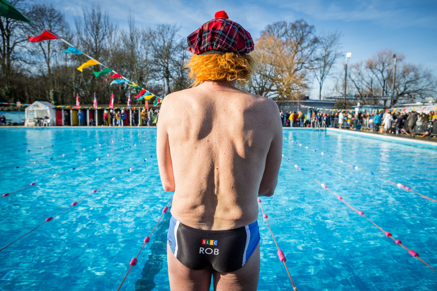 A person with red hair wearing a red plaid hat standing at the edge of an outdoor swimming pool, facing away from the camera. The pool is crowded with people, colorful flags line the pool, and trees are visible in the background.