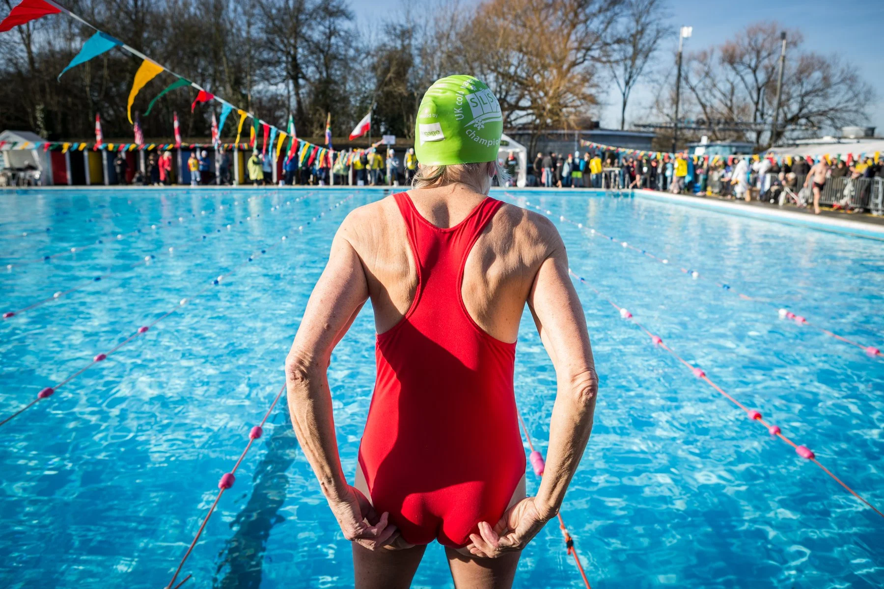 An elderly woman in a red swimsuit and green swim cap standing at the edge of an outdoor swimming pool, preparing to swim. There are spectators behind a barrier and colorful flags strung above the pool area.