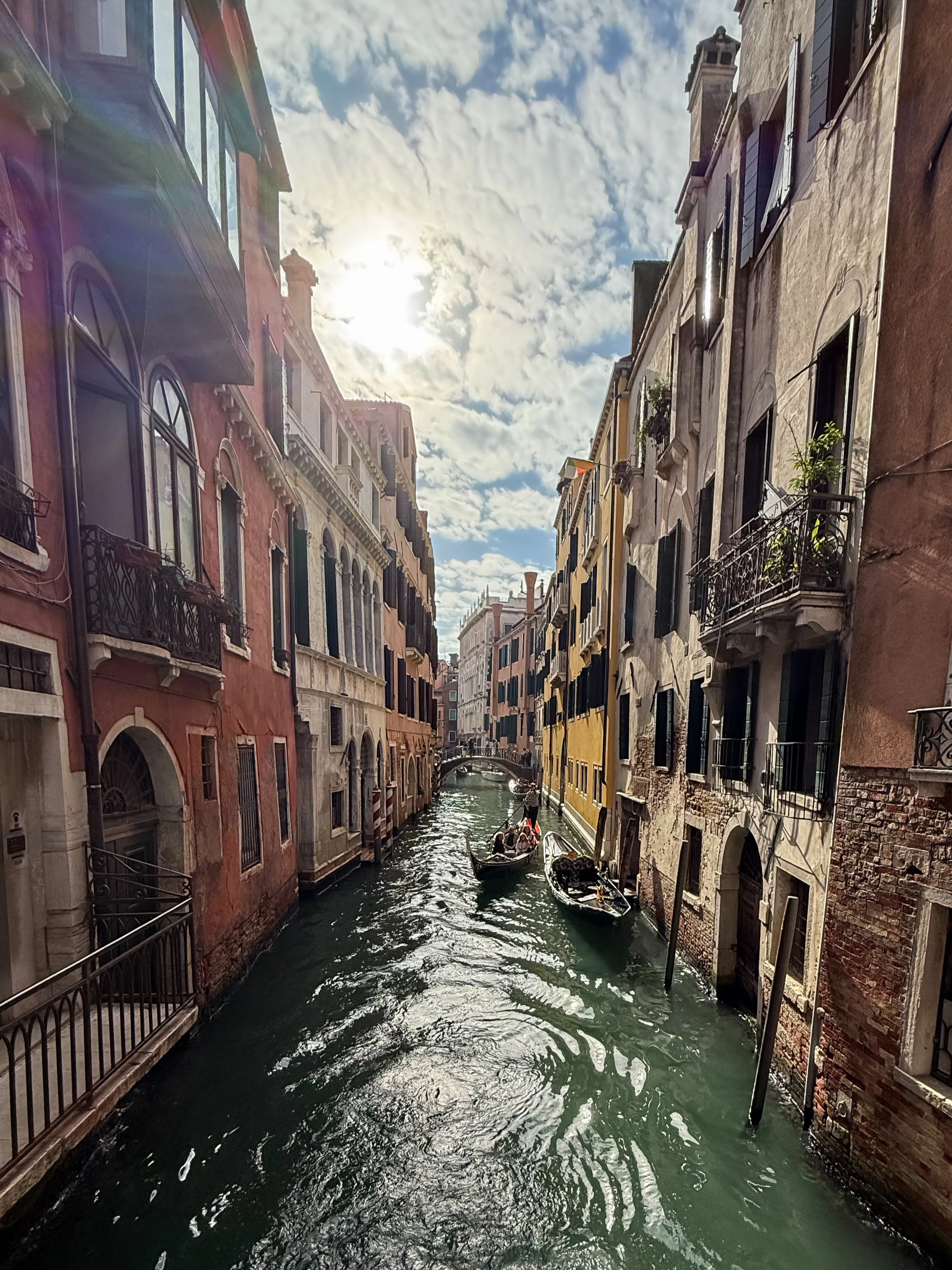 A narrow canal in Venice, Italy with colorful buildings on both sides, a small bridge in the distance, and a boat with passengers navigating the waterway under a partly cloudy sky.
