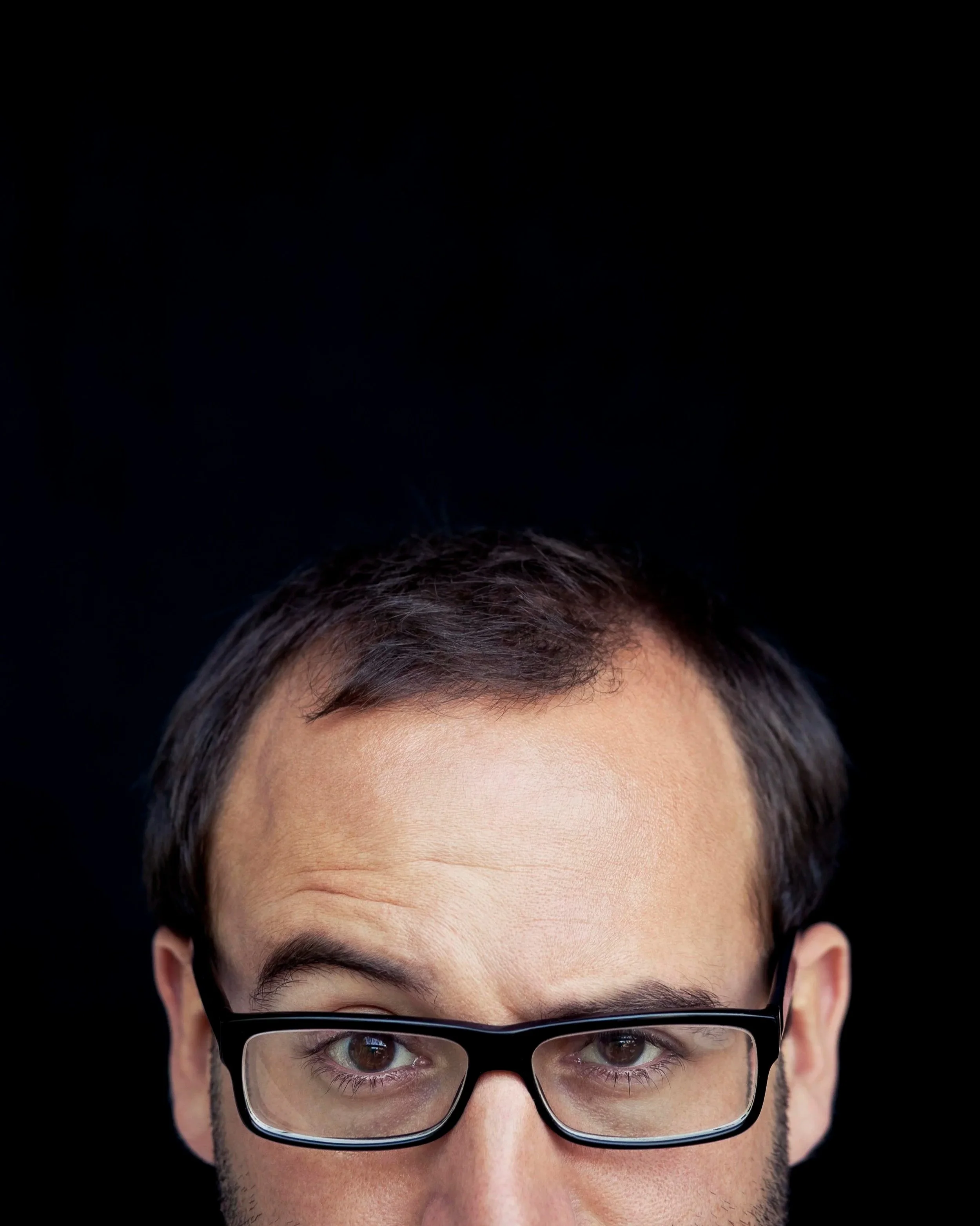 Close-up of a person's forehead with dark hair, eyebrows, and eyes visible through black-rimmed glasses, set against a solid black background.