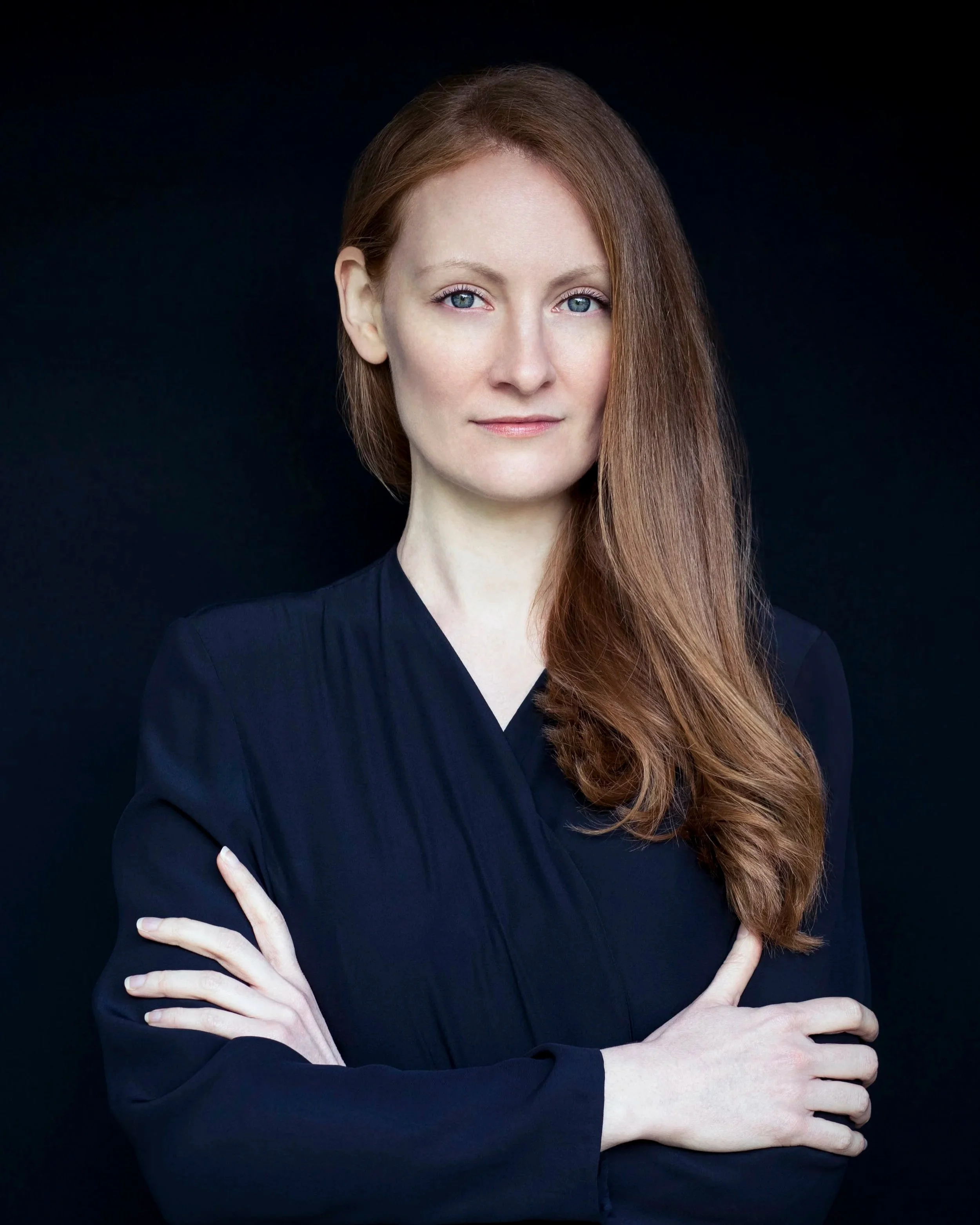 A woman with long red hair and blue eyes wearing a black top, posed with her arms crossed and hands resting on her shoulders, against a black background.