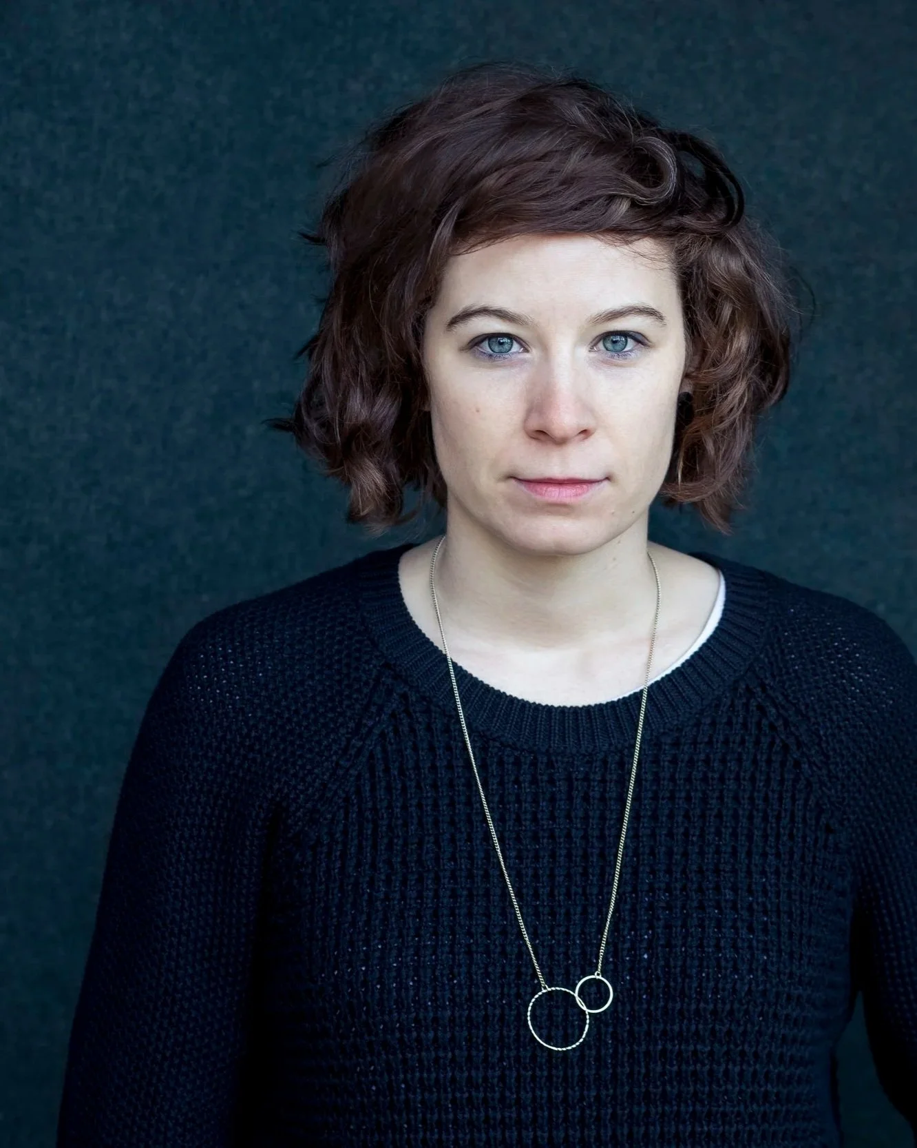 Portrait of a woman with short, wavy brown hair and blue eyes, wearing a black sweater with a silver necklace, standing against a dark background.