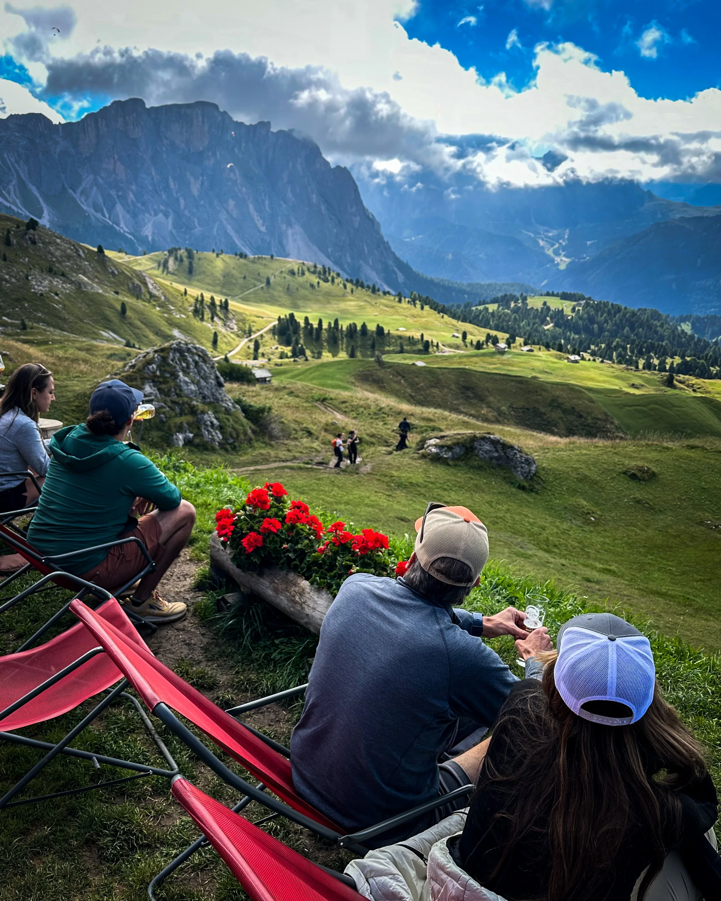 People sitting on chairs and lying on the grass in a scenic mountain landscape, with clear skies, lush green hills, and towering mountains in the background. Some individuals are enjoying drinks and socializing.