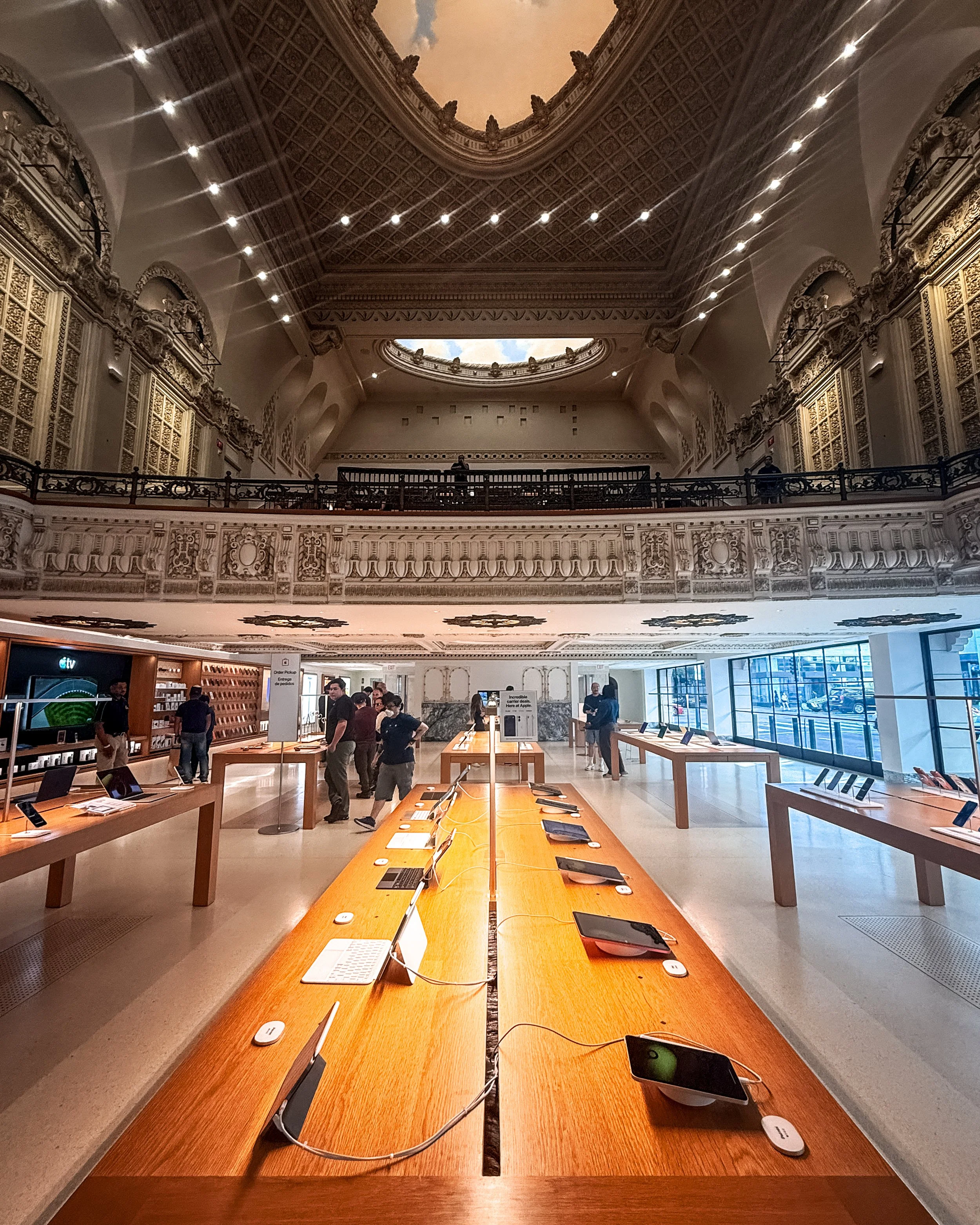 Inside the Tower Theatre Apple Store in Downton Los Angeles, California, USA, with wooden tables displaying iPads, in a spacious high-ceiling retail space with ornate architectural details, large windows on the right.