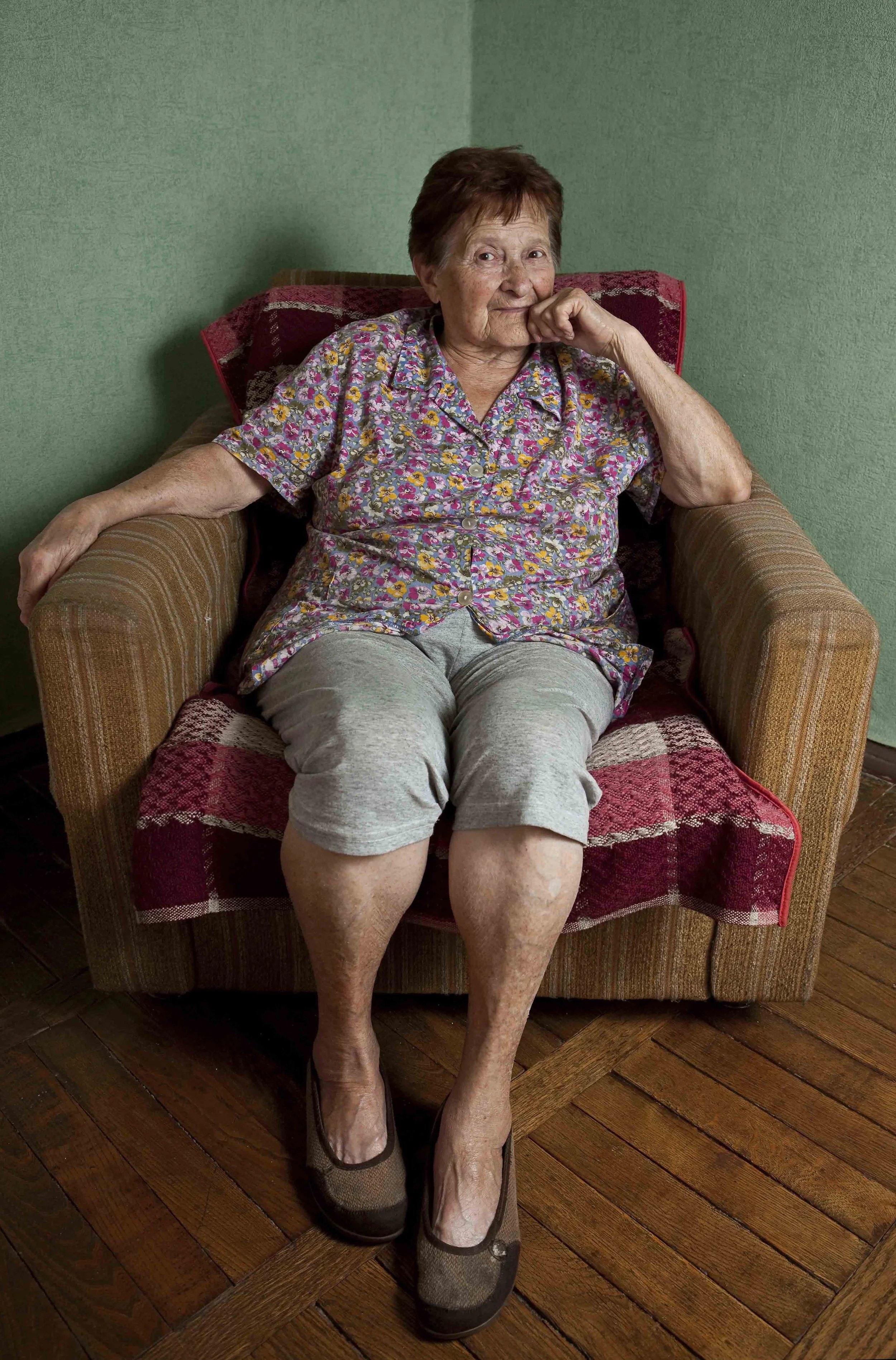 An elderly woman sitting in a brown armchair with a red and pink patterned blanket on the backrest. She is wearing a floral short-sleeve shirt, gray shorts, and brownish-gray sandals. She has short brown hair and is sitting against a green wall with 