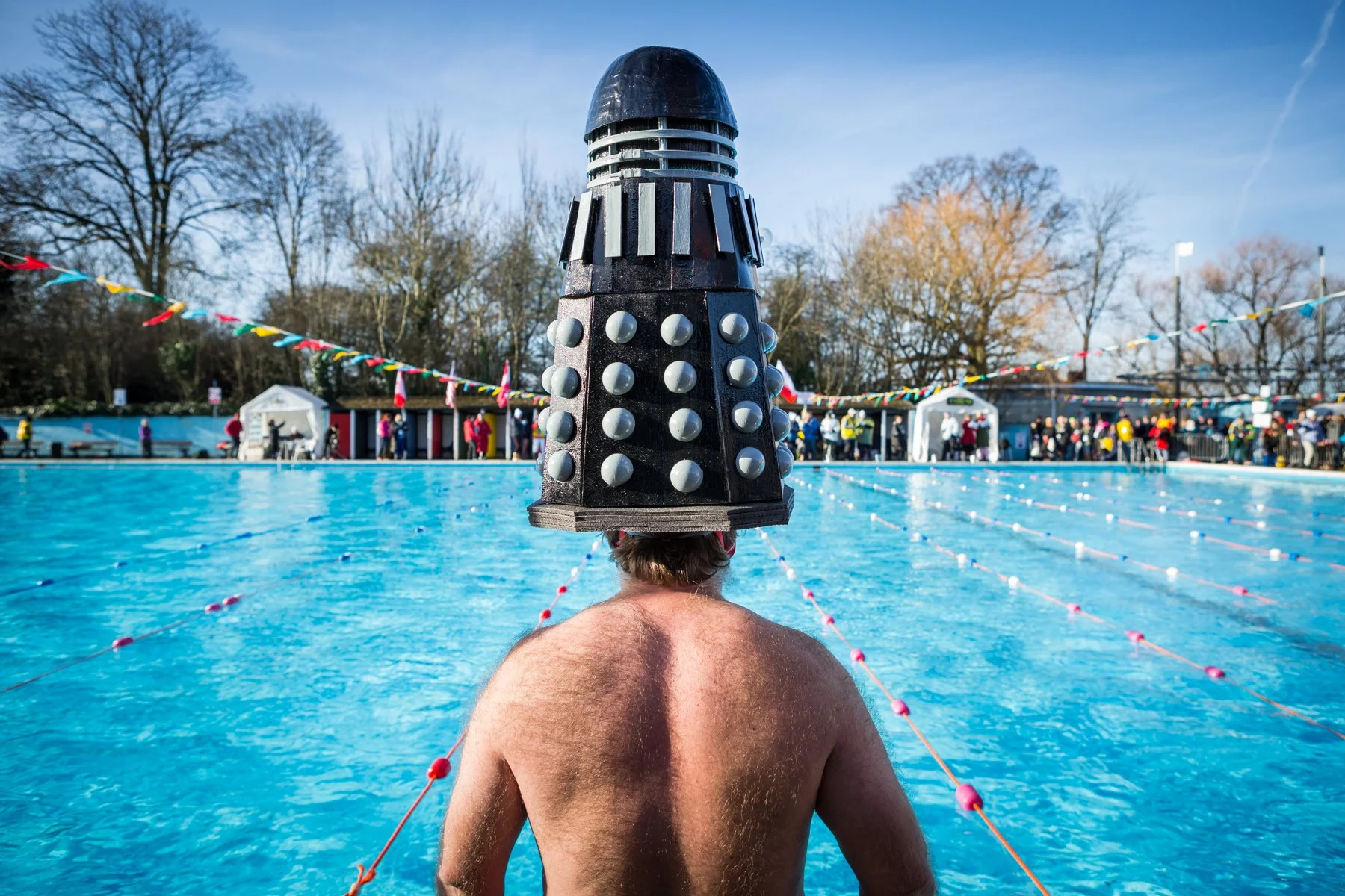 A person standing in a swimming pool with their back to the camera, wearing a homemade Dalek costume on their head, during an event with many spectators and colorful bunting.