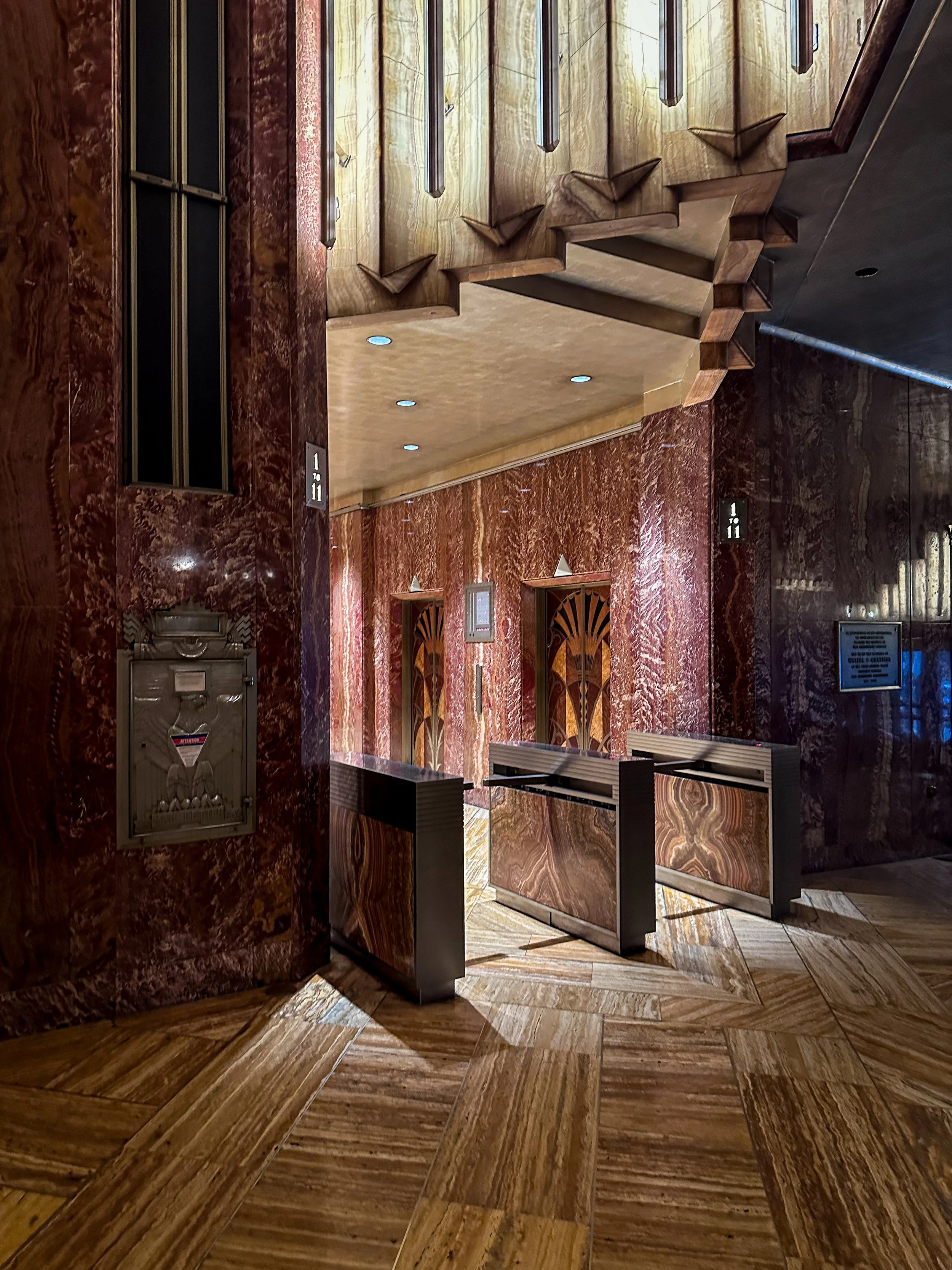 Interior of a building with marble walls and floors, an elevator door, and a staircase with wooden steps and a decorative railing.