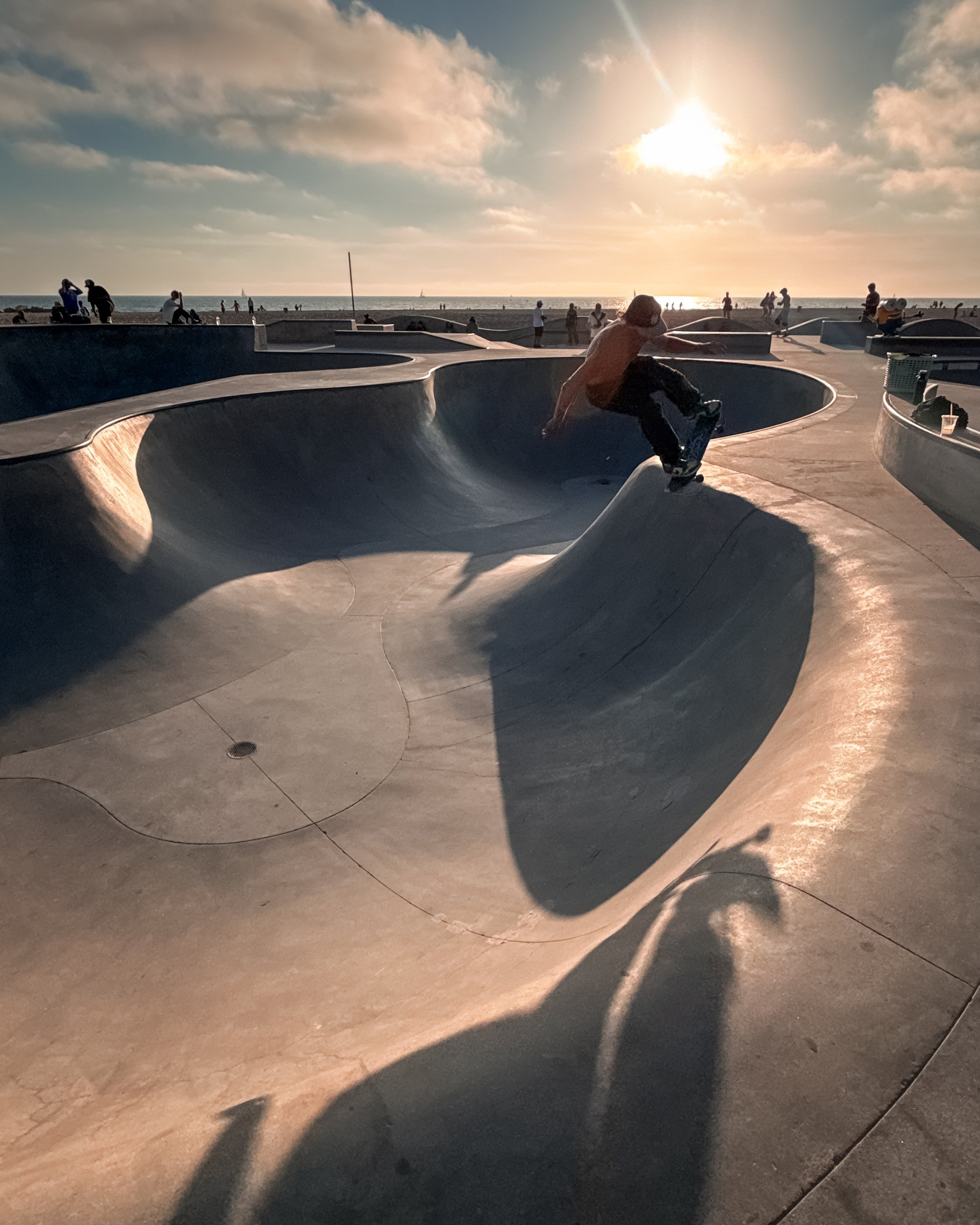 The skatepark at Venice Beach, Los Angeles, California, USA, with a person skating on a curved, smooth concrete bowl during sunset. Several people are standing and sitting around the skatepark, and the ocean is visible in the background