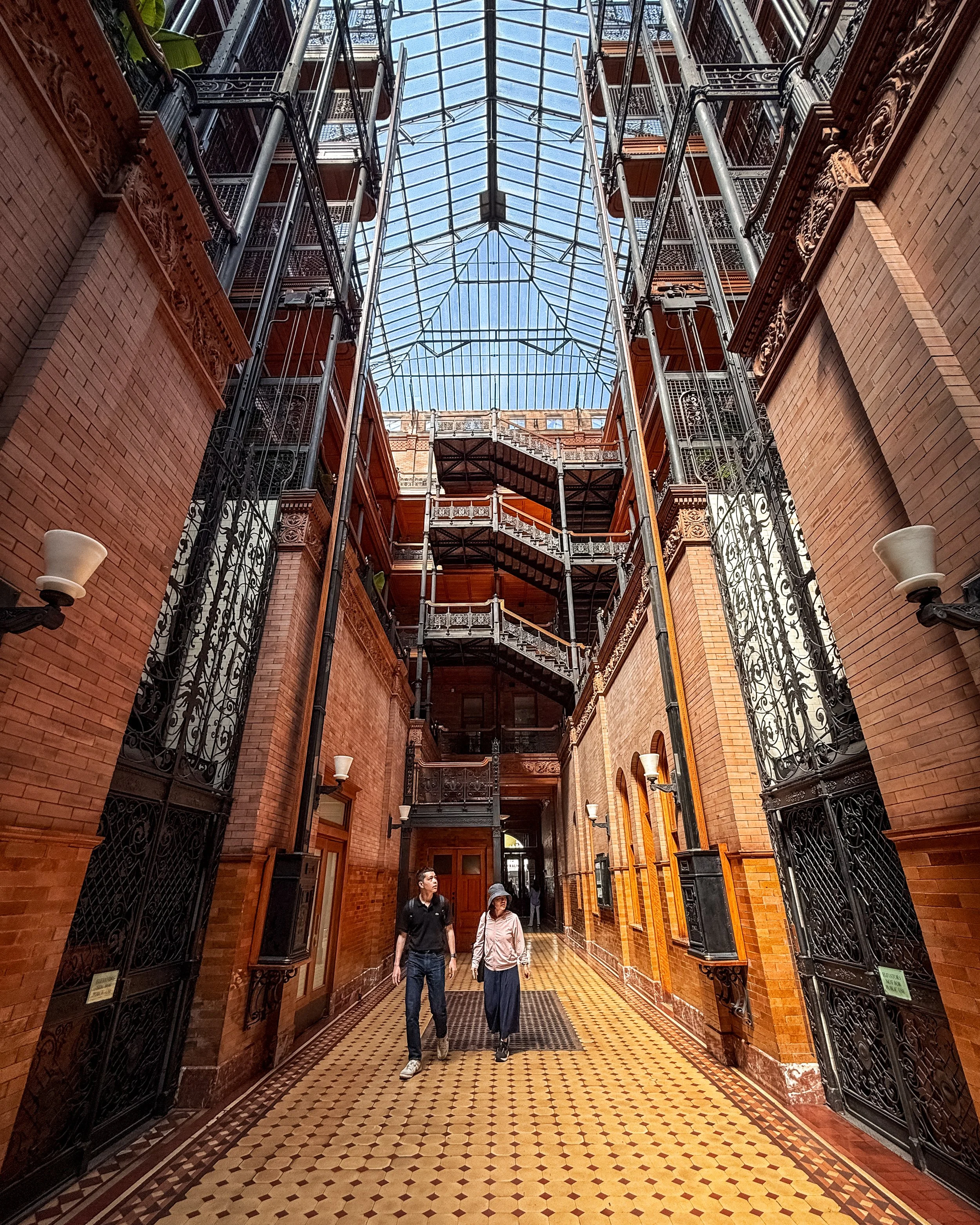 Interior view of the Bradbury Building in Downton Los Angeles, California, USA, with a glass ceiling, ornate ironwork, brick walls, and patterned tile floor. Two people are walking in the corridor.