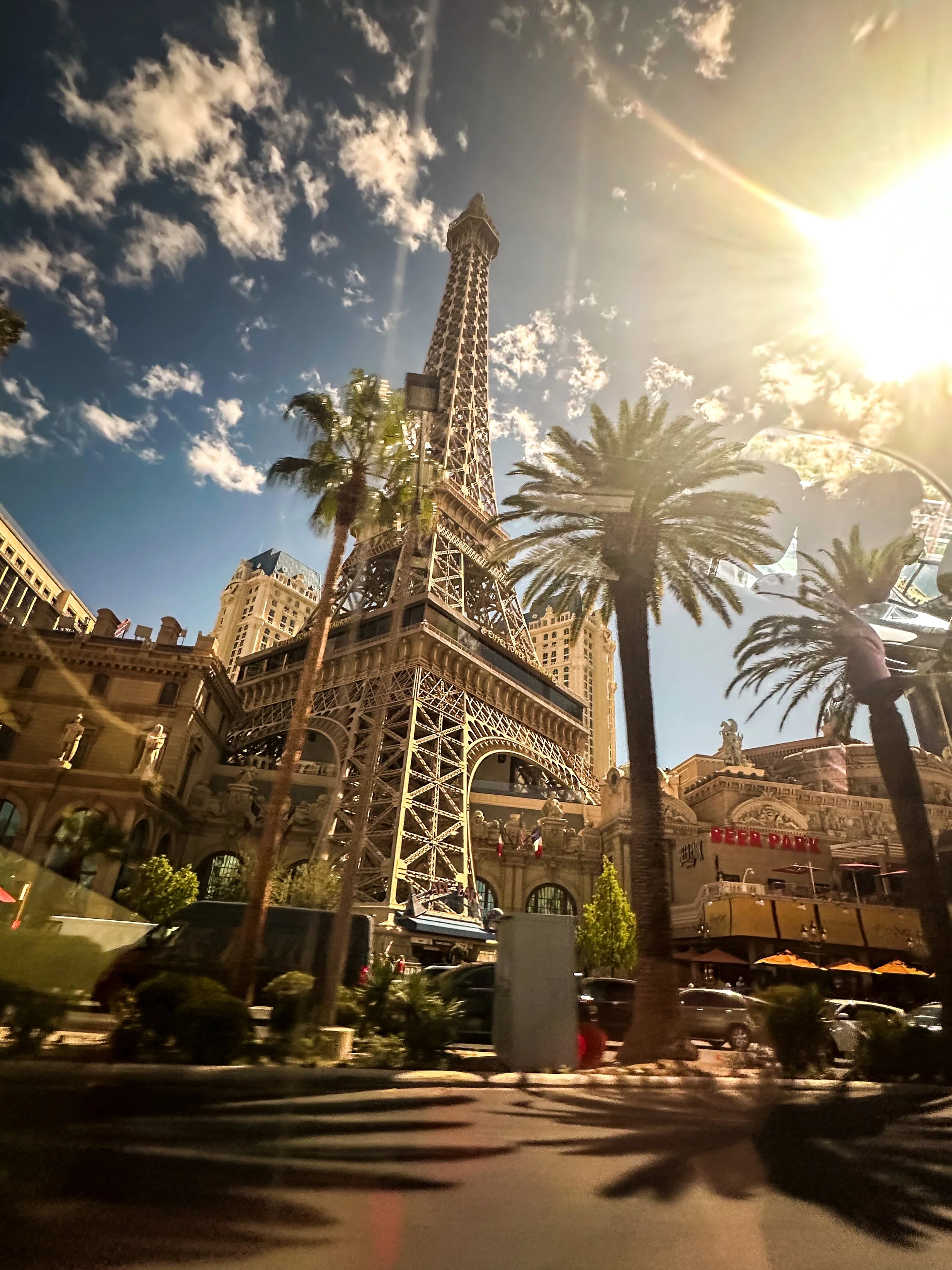 Tall replica of the Eiffel Tower with palm trees in front, and a backdrop of buildings under a partly cloudy sky with the sun shining brightly from the right side.