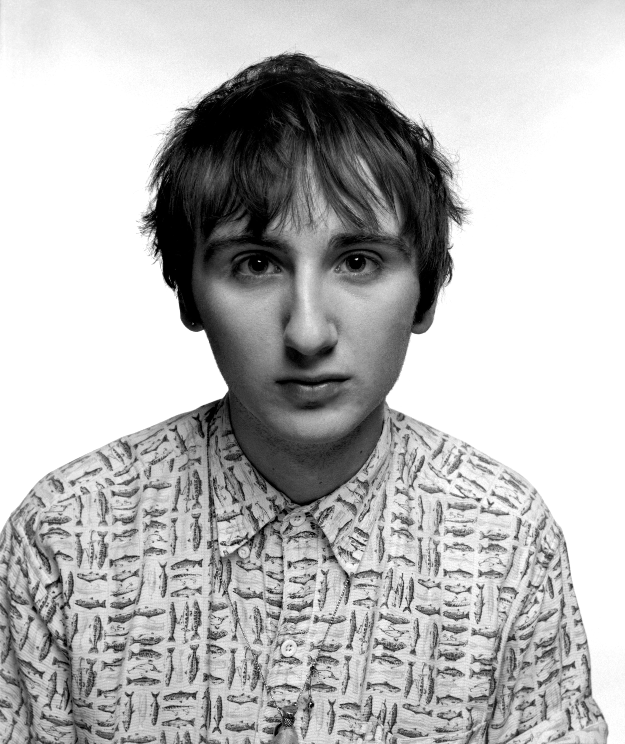 Close-up portrait of a young man with short, messy hair, wearing a shirt and a necklace. Medium format film photograph.