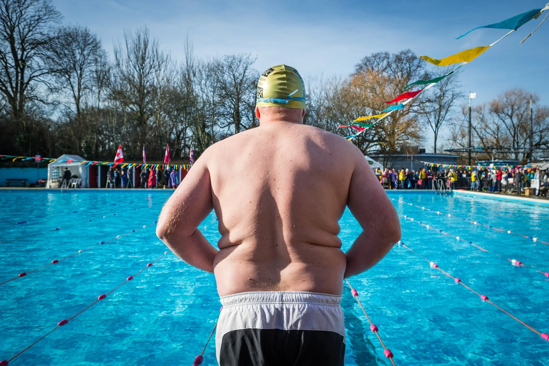 An elderly man wearing a yellow swim cap and swim trunks stands at the edge of an outdoor swimming pool, preparing for a swim. The pool is filled with blue water and is lined with ropes and colorful flags, with spectators and flags visible in the bac