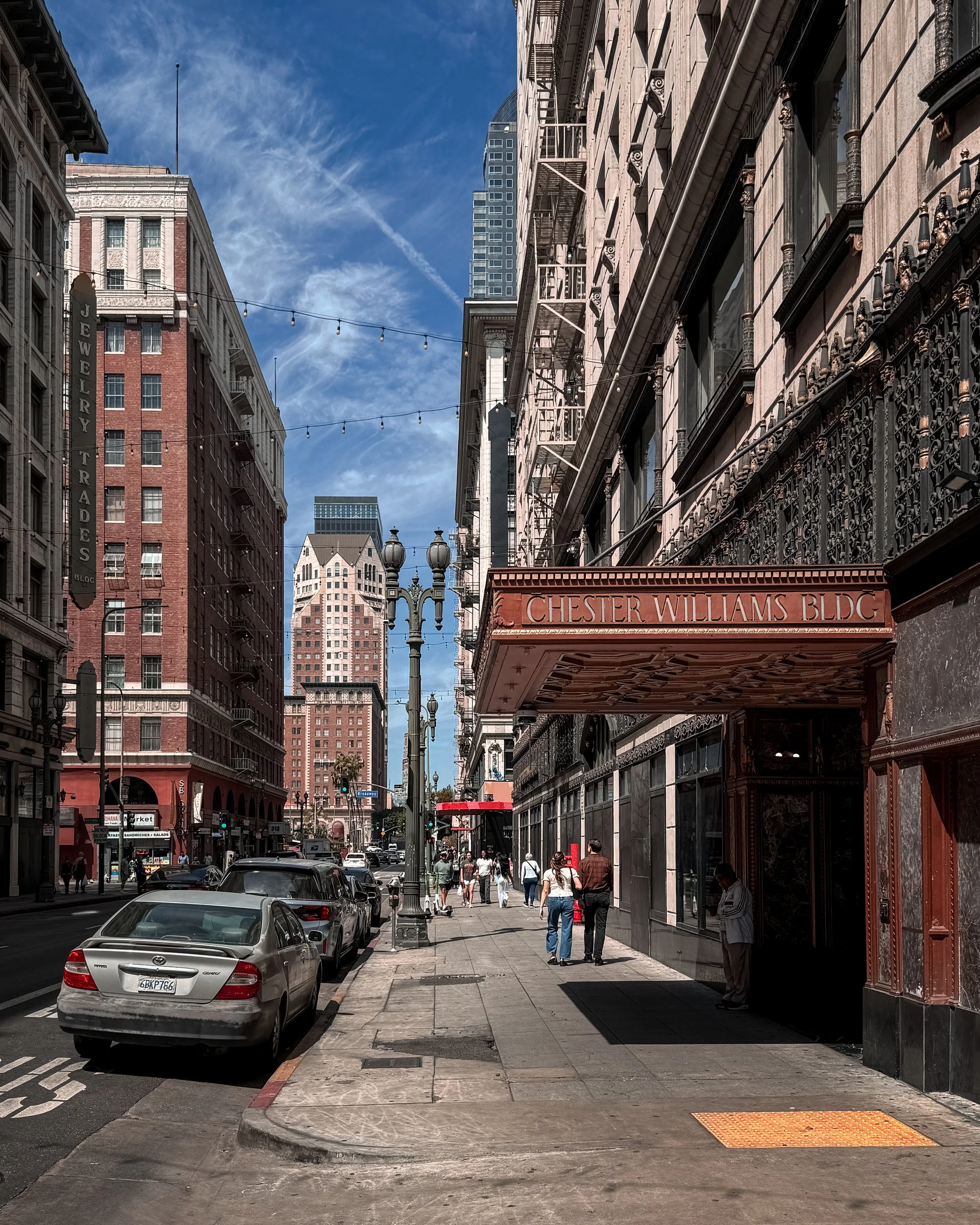 A sunny street scene in Downton Los Angeles, California, USA, with parked cars on the left and pedestrians walking on the sidewalk. Tall buildings with ornate architectural details line the street.
