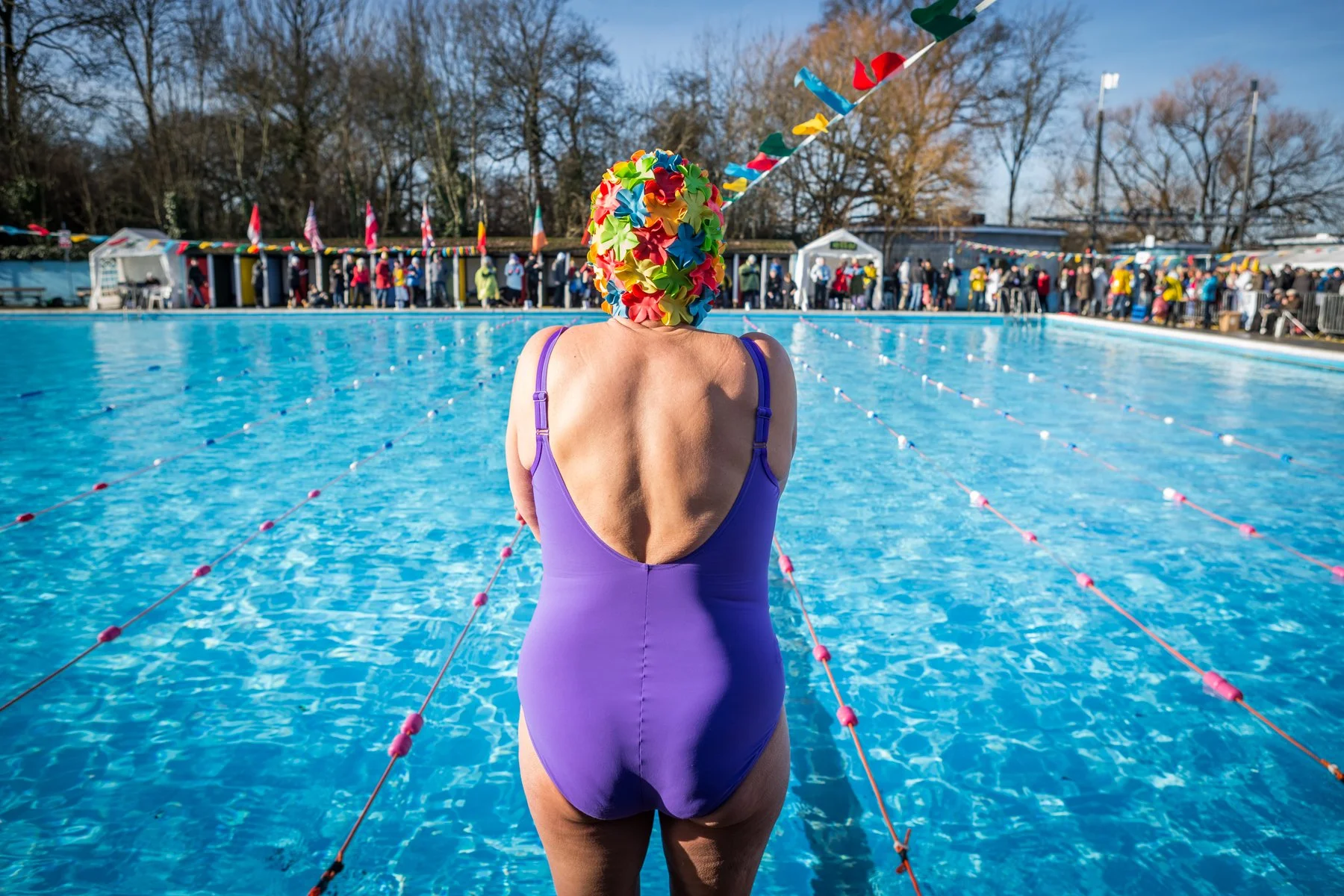 A woman in a purple swimsuit and a colorful floral swim cap stands at the edge of a swimming pool, facing away. The pool is surrounded by people, with some tents and trees in the background on a clear, sunny day.