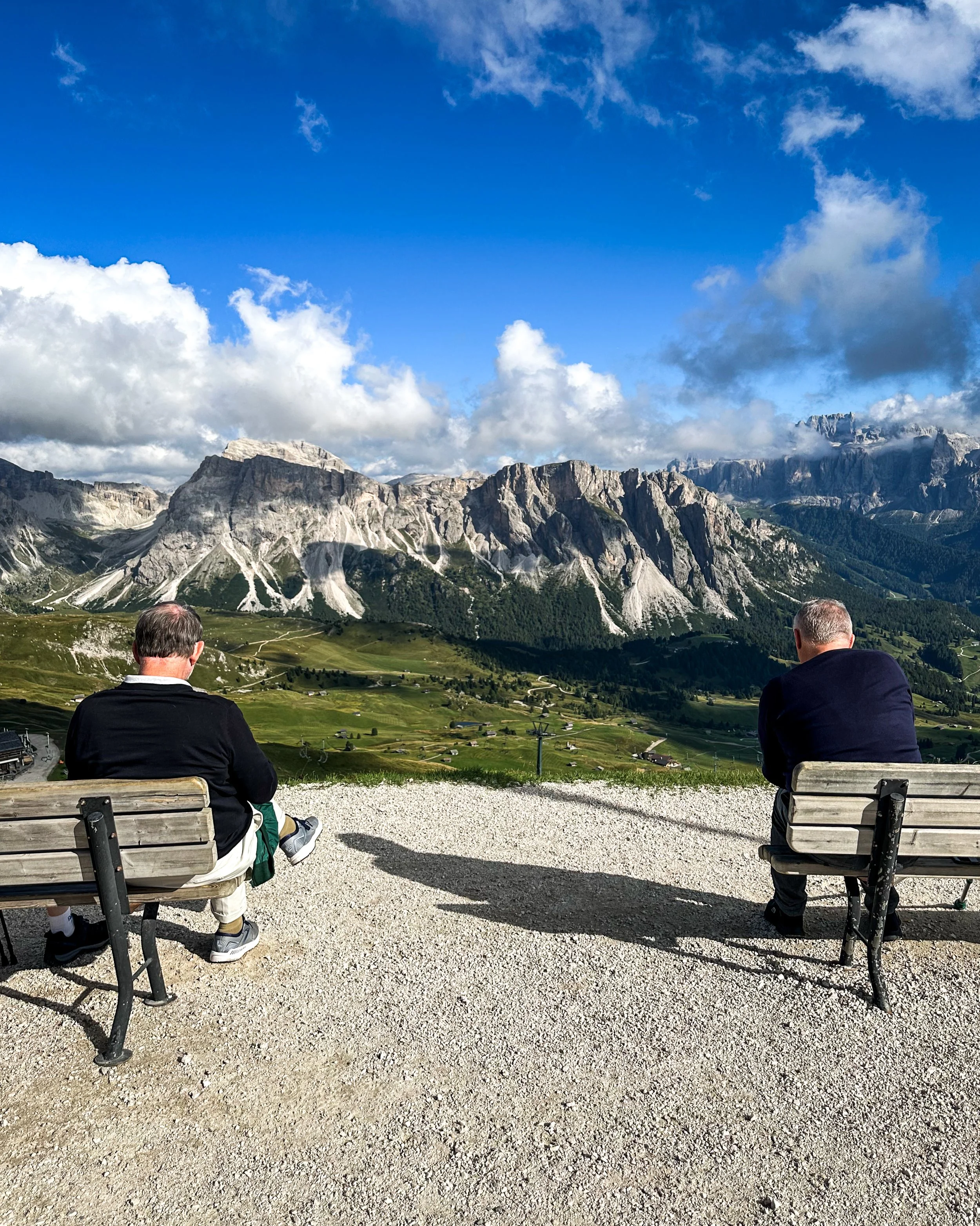 Two men sitting on wooden benches at a scenic mountain viewpoint under a partly cloudy sky, with a view of green fields, mountains, and rocky peaks in the background.