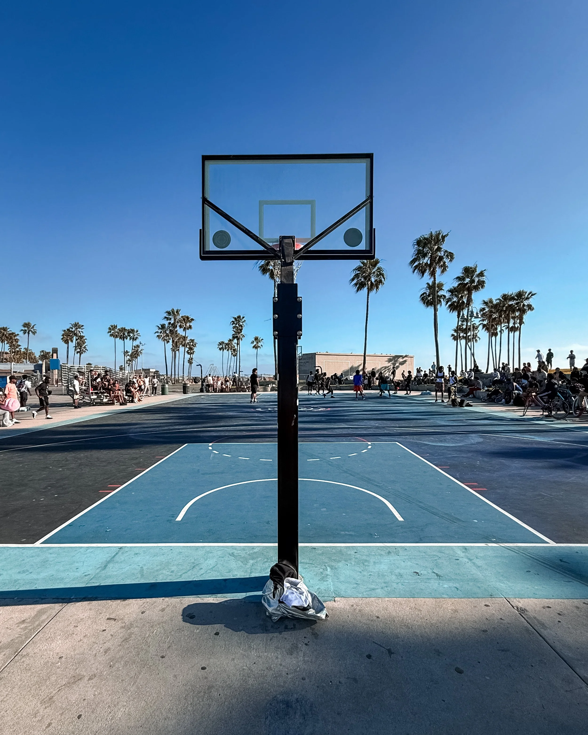 A view from behind a basketball hoop on an outdoor court, with a bag placed on the ground in front. The court is crowded with people playing and relaxing, and there are tall palm trees under a clear blue sky in the background.