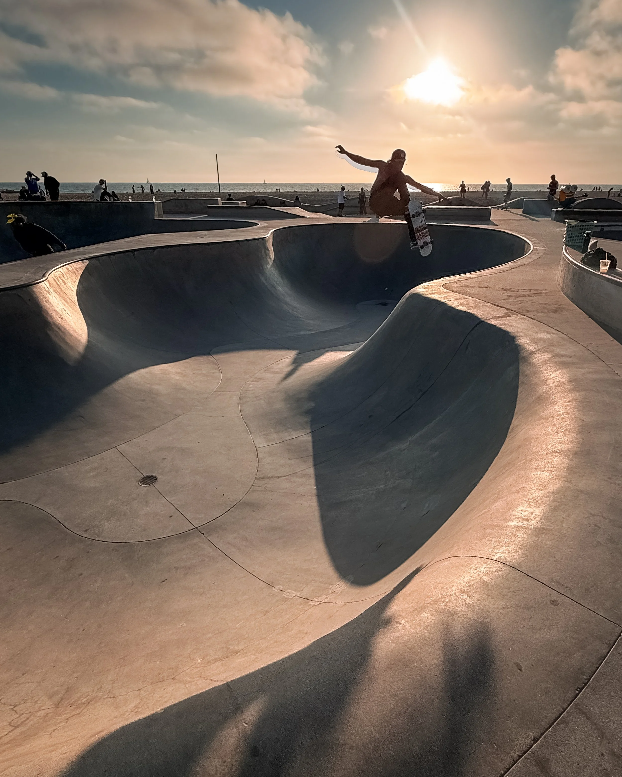 The skatepark at Venice Beach, Los Angeles, California, USA, with a person skating on a curved, smooth concrete bowl during sunset. Several people are standing and sitting around the skatepark, and the ocean is visible in the background