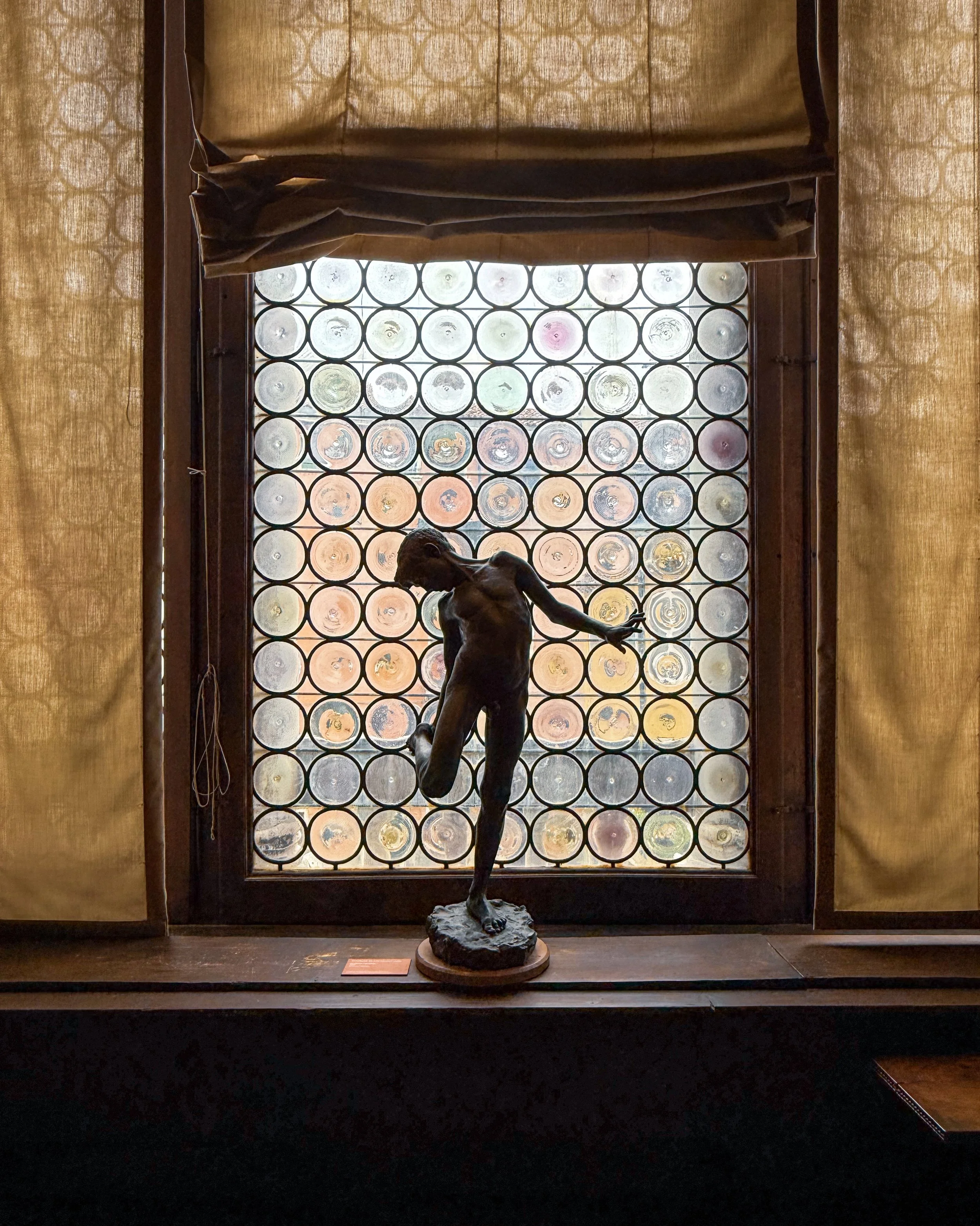 Statue of a woman with an elegant pose, standing on a rock-like base, placed in front of a glass window with circular patterns, inside a museum display.