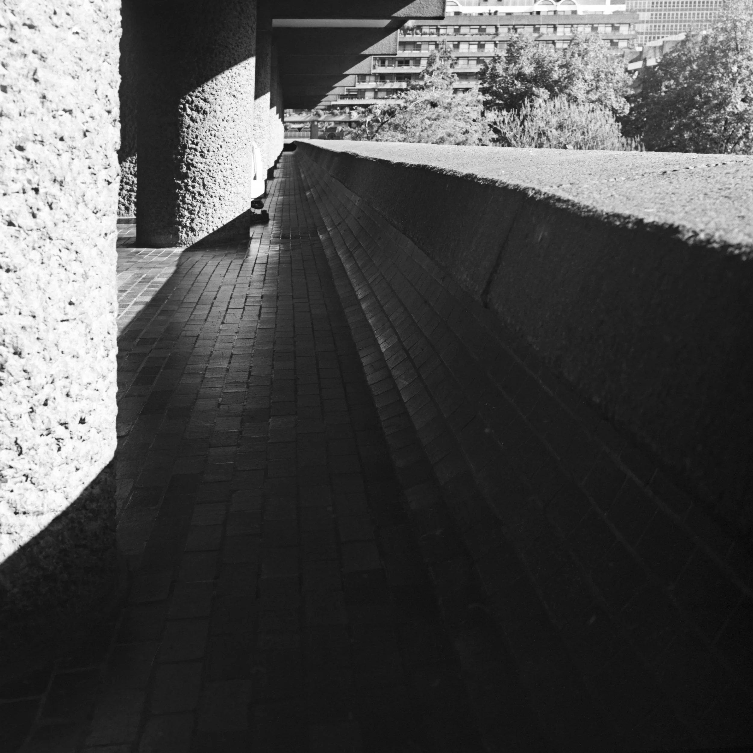 Black and white photo taken from a terrace in London, United Kingdom, showing Brutalist textured stone columns, a sloped tiled roof or ledge, and modern high-rise buildings and trees in the background.