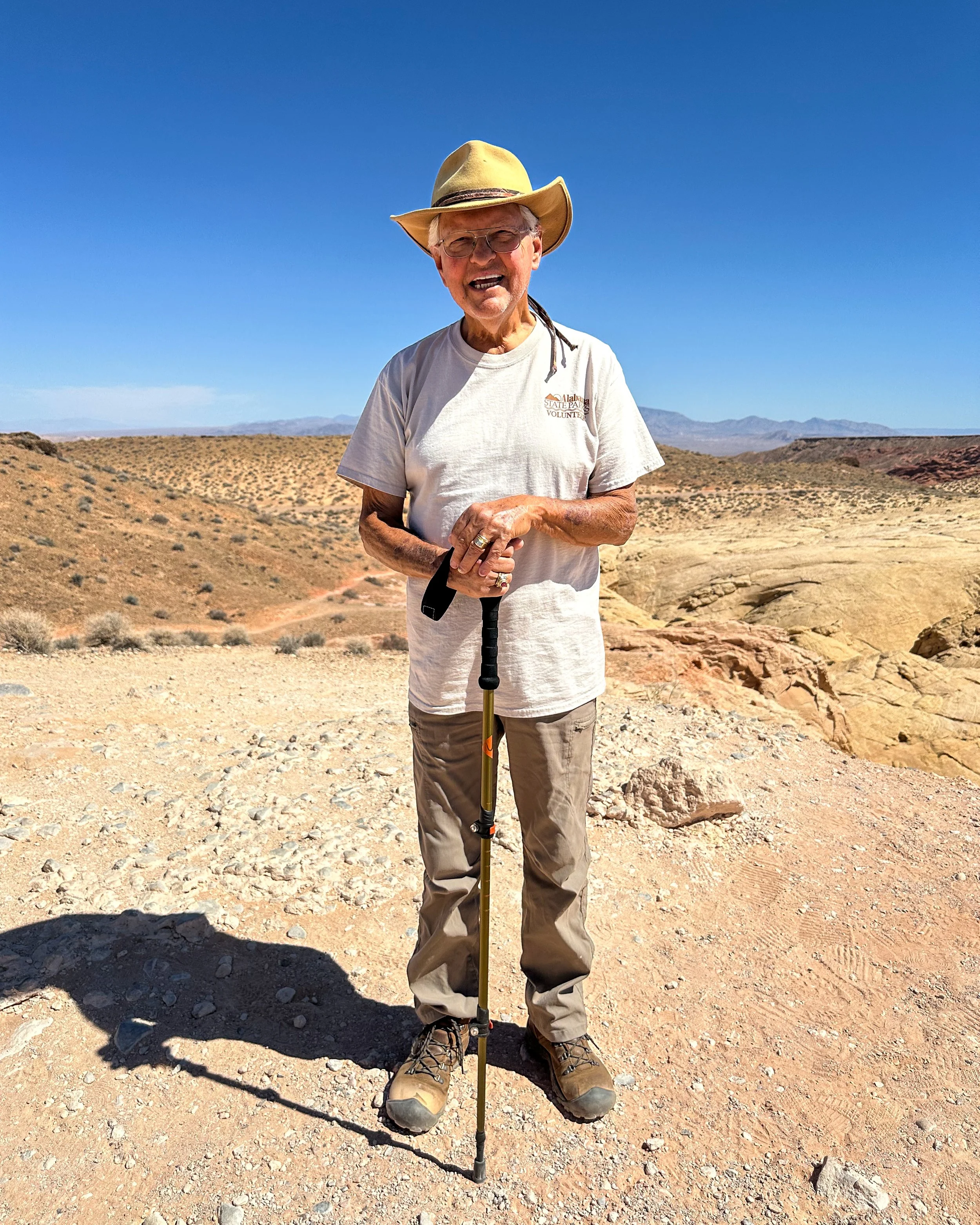 An elderly man standing in a desert landscape, wearing a wide-brimmed hat, glasses, a white T-shirt, and tan hiking boots. He is smiling and holding a trekking pole. The background features mountains and a clear blue sky.