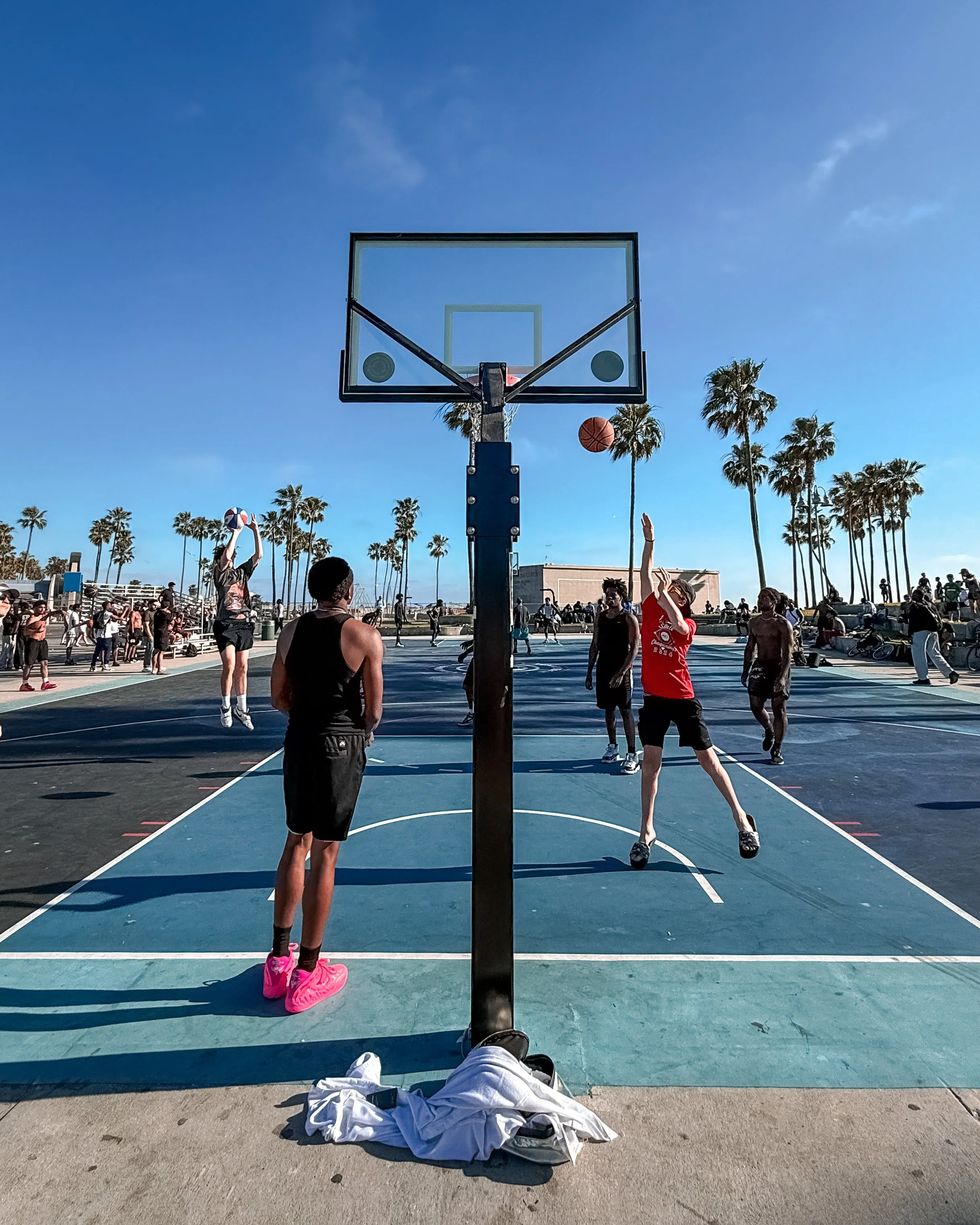 People playing basketball on an outdoor court with palm trees in the background, taken from the perspective of a person sitting on the ground with their legs outstretched.
