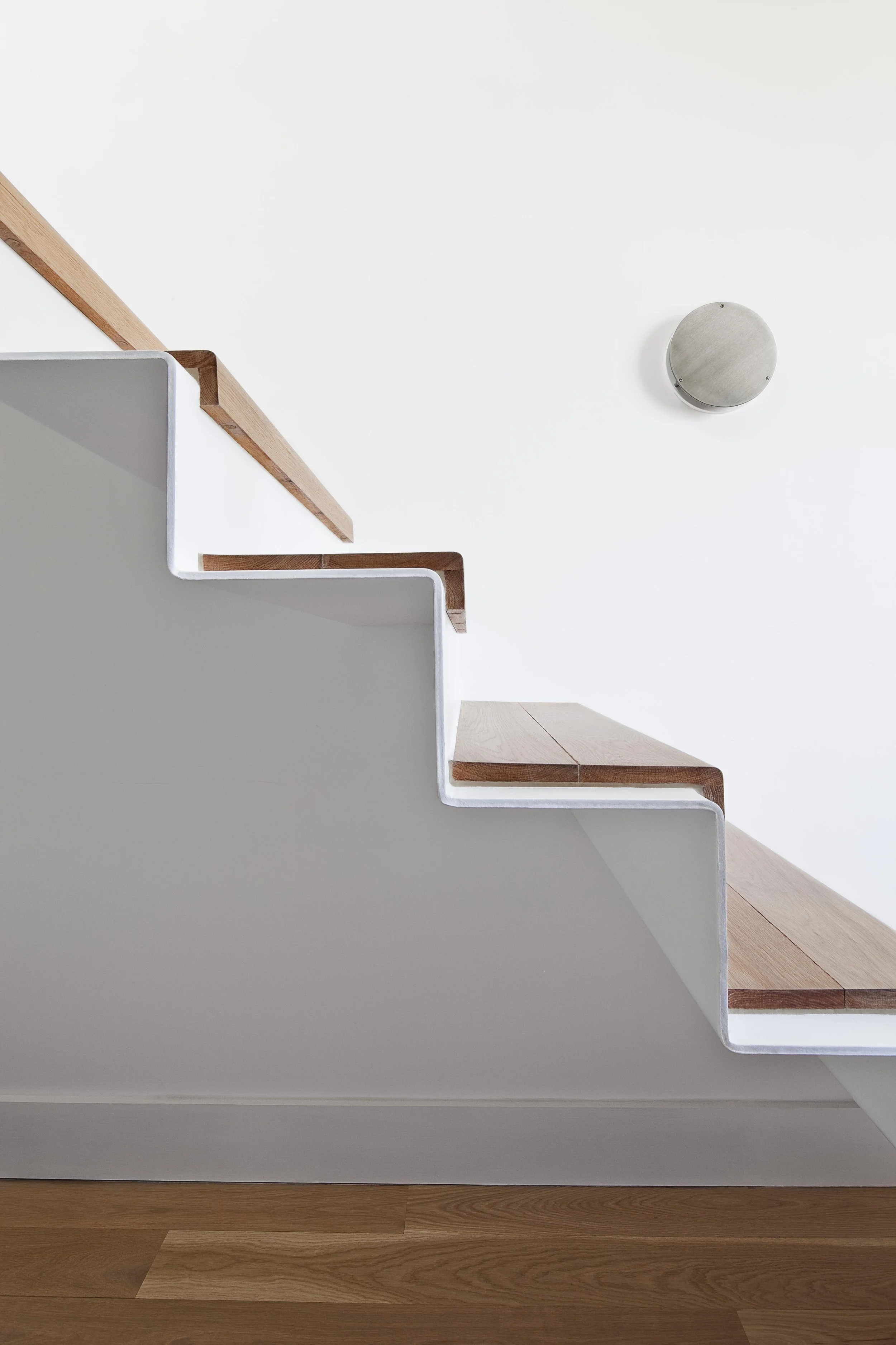 Interior view of a staircase with wooden steps and white sides, seen from below, with a white wall and a circular metallic light fixture on the ceiling.