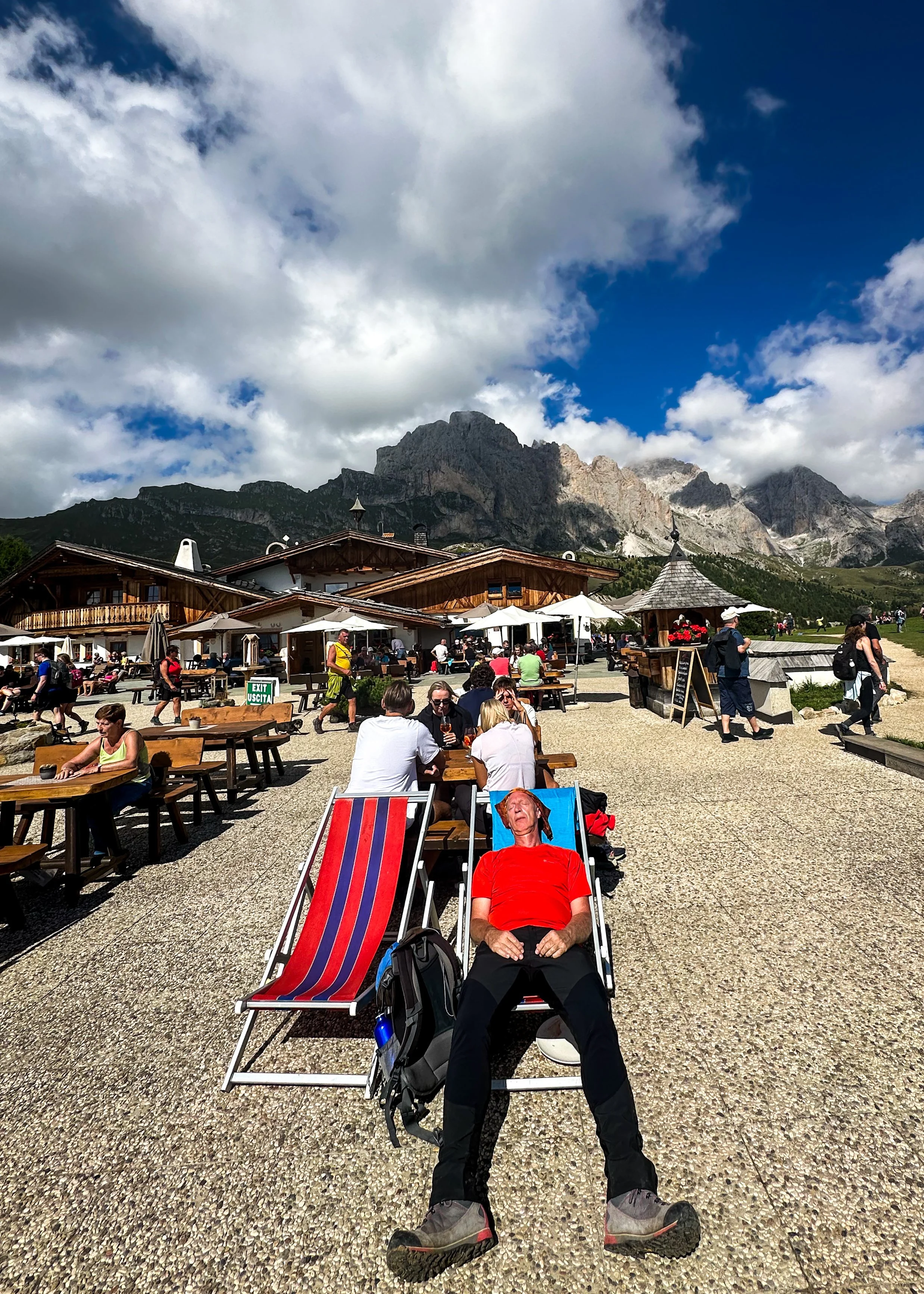 A man in red shirt and black pants relaxing on a deck chair with red and blue stripes, outdoors on a sunny day with mountains in the background, surrounded by other people sitting at tables and walking.