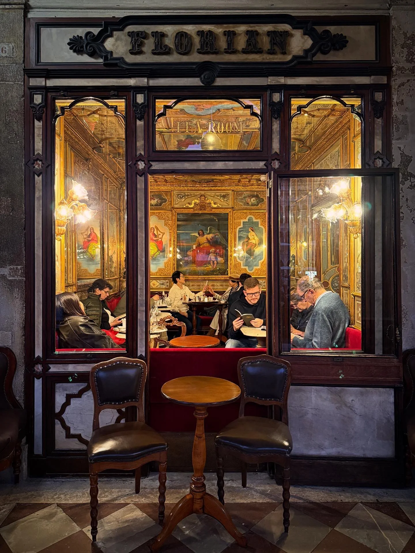 A view through a large window of a restaurant cafe tea room called Florian in Venice, Italy, with people dining inside and ornate decorations on the walls.