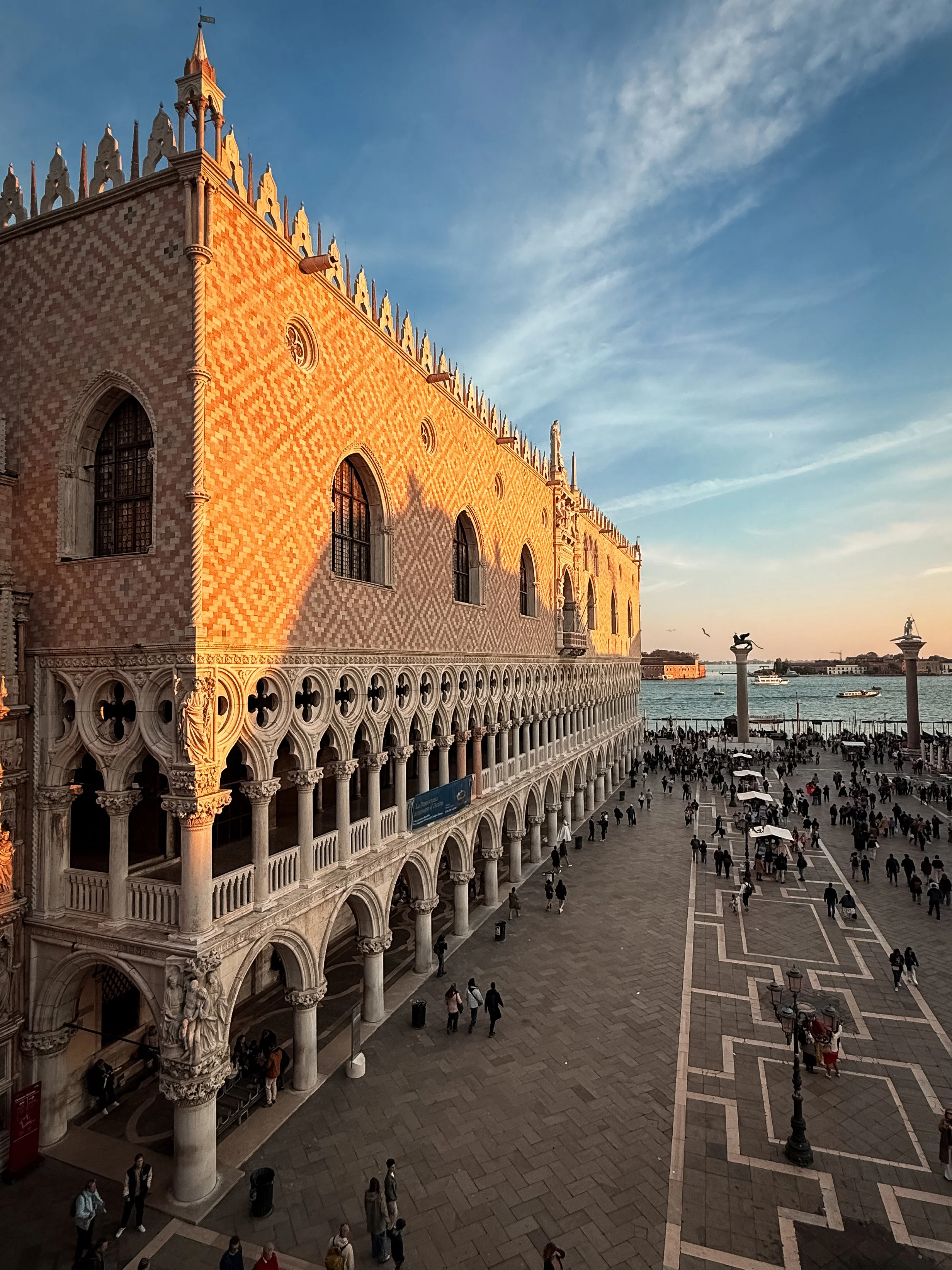 Doge's Palace or Palazzo Ducale in Venice, Italy, with intricate architecture, arched windows, and statues, overlooking the Grand Canal with boats, and a large open plaza filled with people, during sunset. As seen from Saint Mark's Basilica.