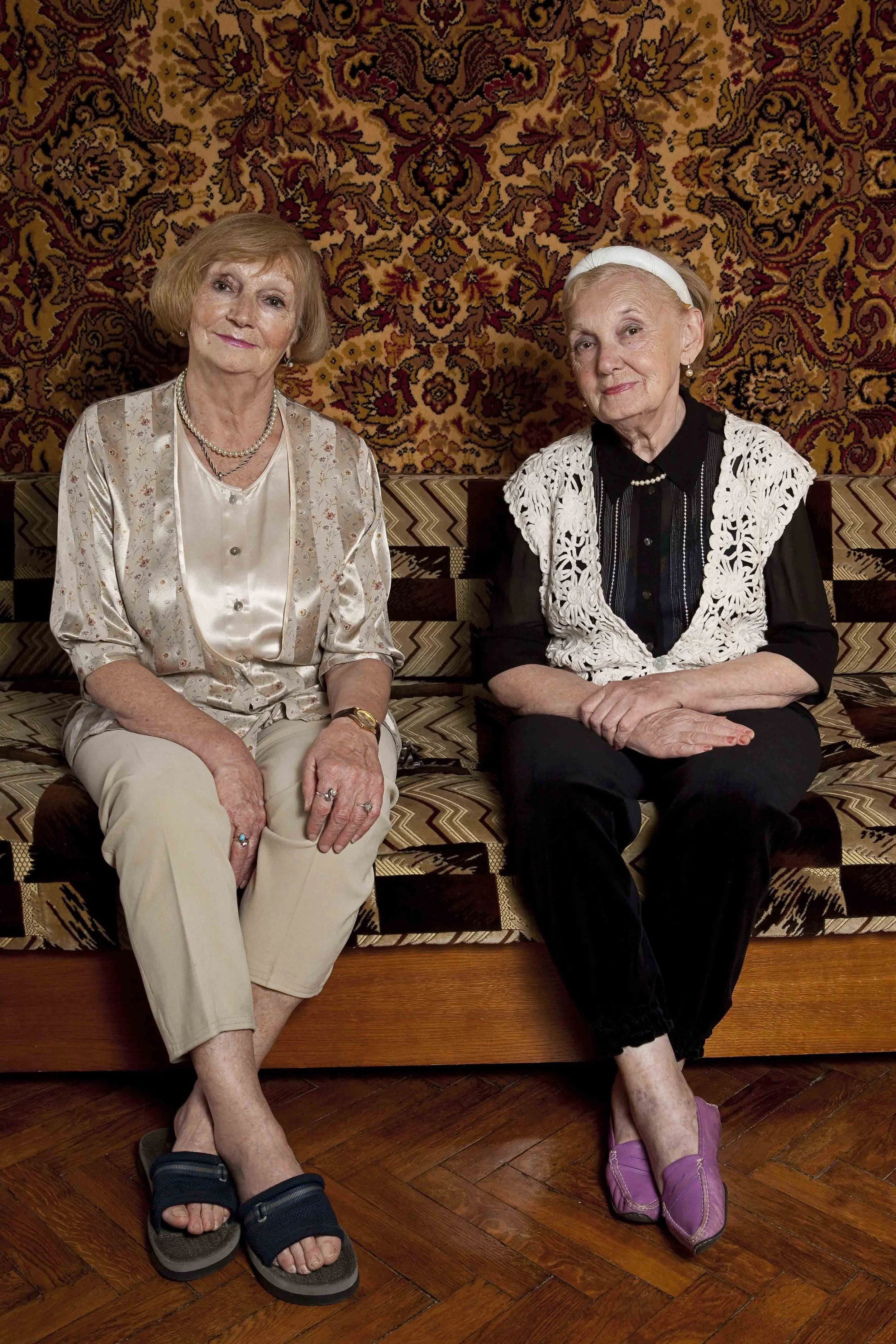 Two elderly women sitting on a sofa in front of a patterned tapestry, one wearing a beige satin blouse and cream pants, the other in a black dress with a white crochet vest and purple shoes.