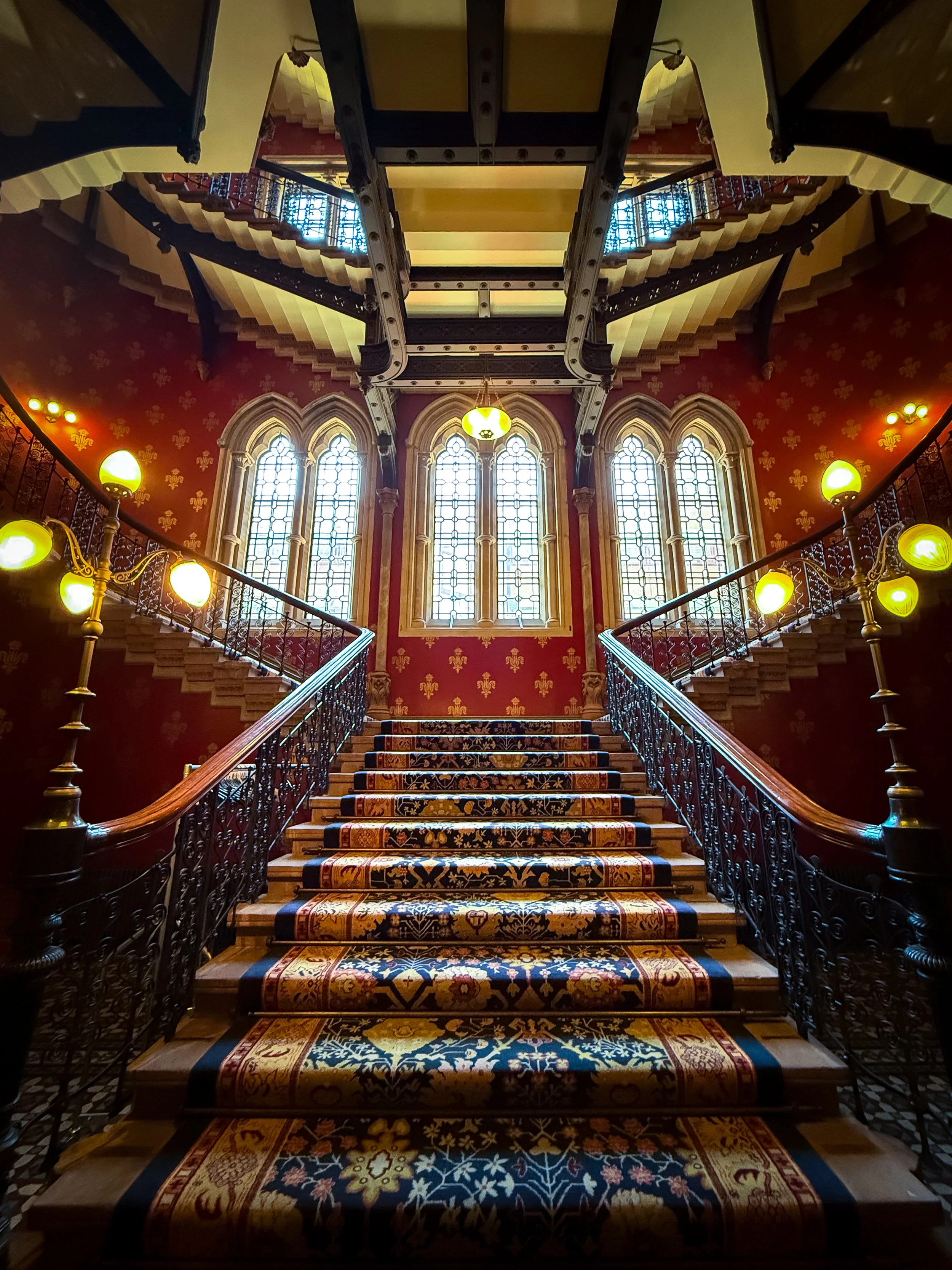 Interior view of the grand staircase in the St Pancras Renaissance Hotel in London, United Kingdom, featuring ornate black iron railings, a colourful patterned carpet on the steps, large stained glass windows.
